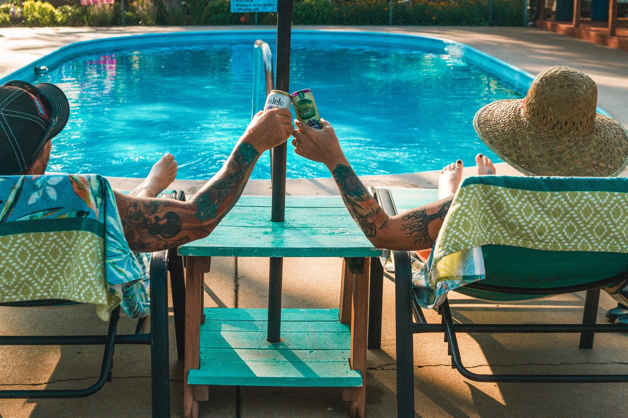 Two people with tattoos sitting by a swimming pool, raising glasses of drinks, one wearing a straw hat.