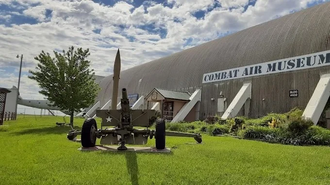 An outdoor display at the Combat Air Museum featuring a large artillery piece, an aircraft, and a military building with a sign reading 'Combat Air Museum' under a partly cloudy sky.