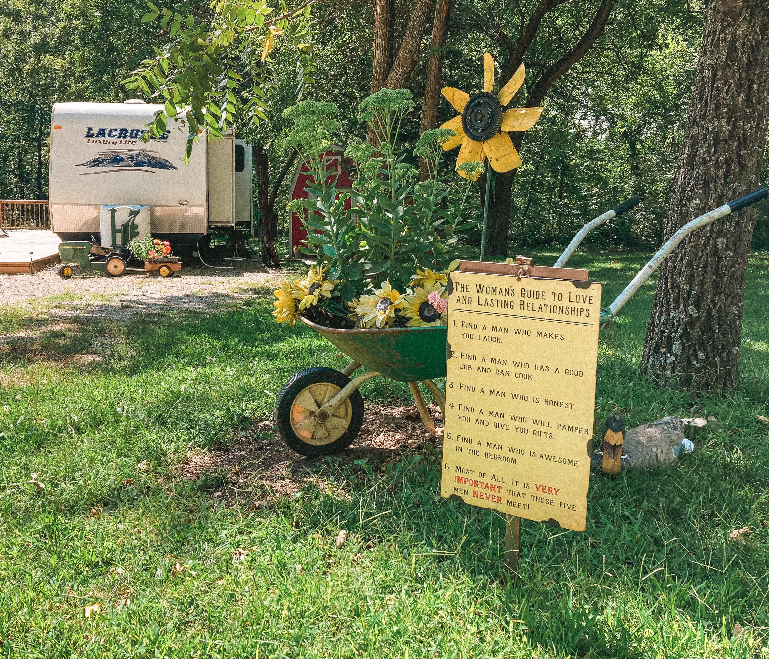 A decorative garden arrangement with a green wheelbarrow filled with yellow and pink flowers, a large sunflower sculpture, and a sign titled "The Woman's Guide to Love and Lasting Relationships" listing five qualities of men, placed outdoors on the g