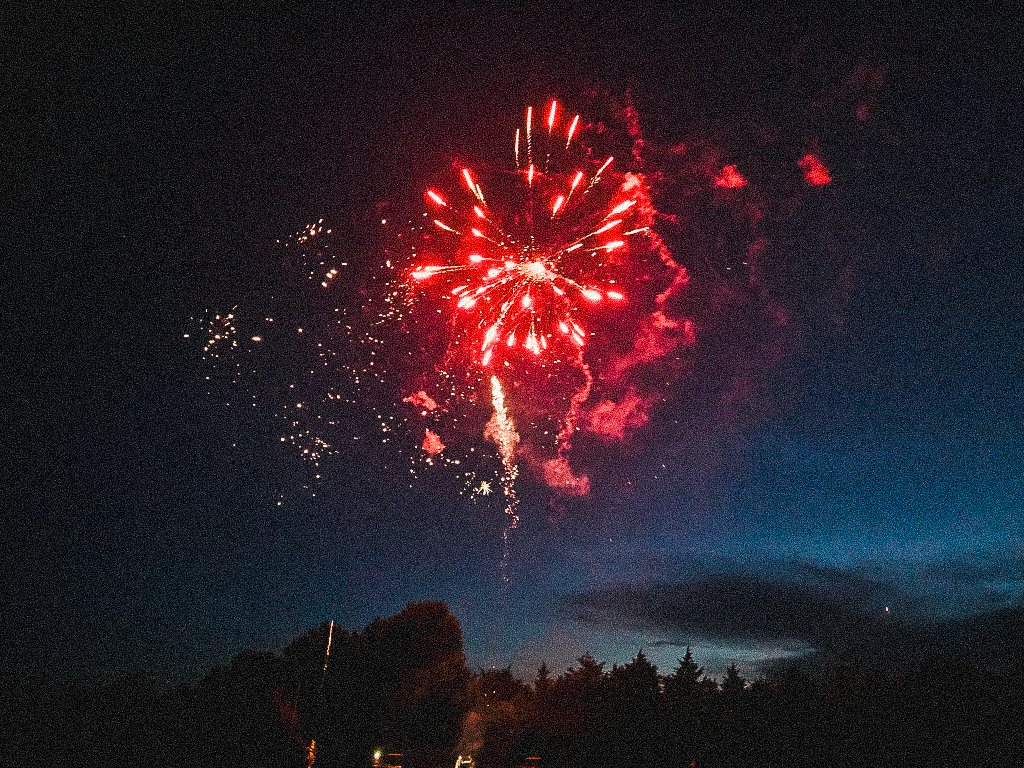 Red fireworks exploding in the night sky above silhouetted trees and clouds.