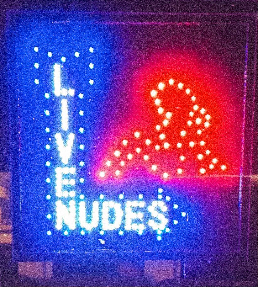 Bright neon sign with the words "Lick Nudes" in colorful lights, featuring a prominent arrow pointing down.