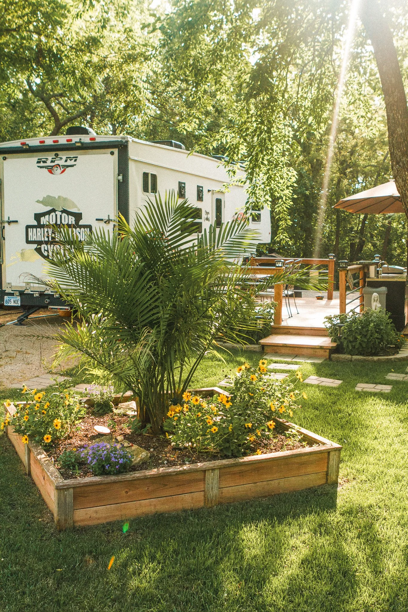 A backyard garden with a raised wooden flower bed containing a large palm and yellow and purple flowers, a grassy lawn, a wooden deck with outdoor furniture, and a white RV with Harley-Davidson graphics parked among trees.