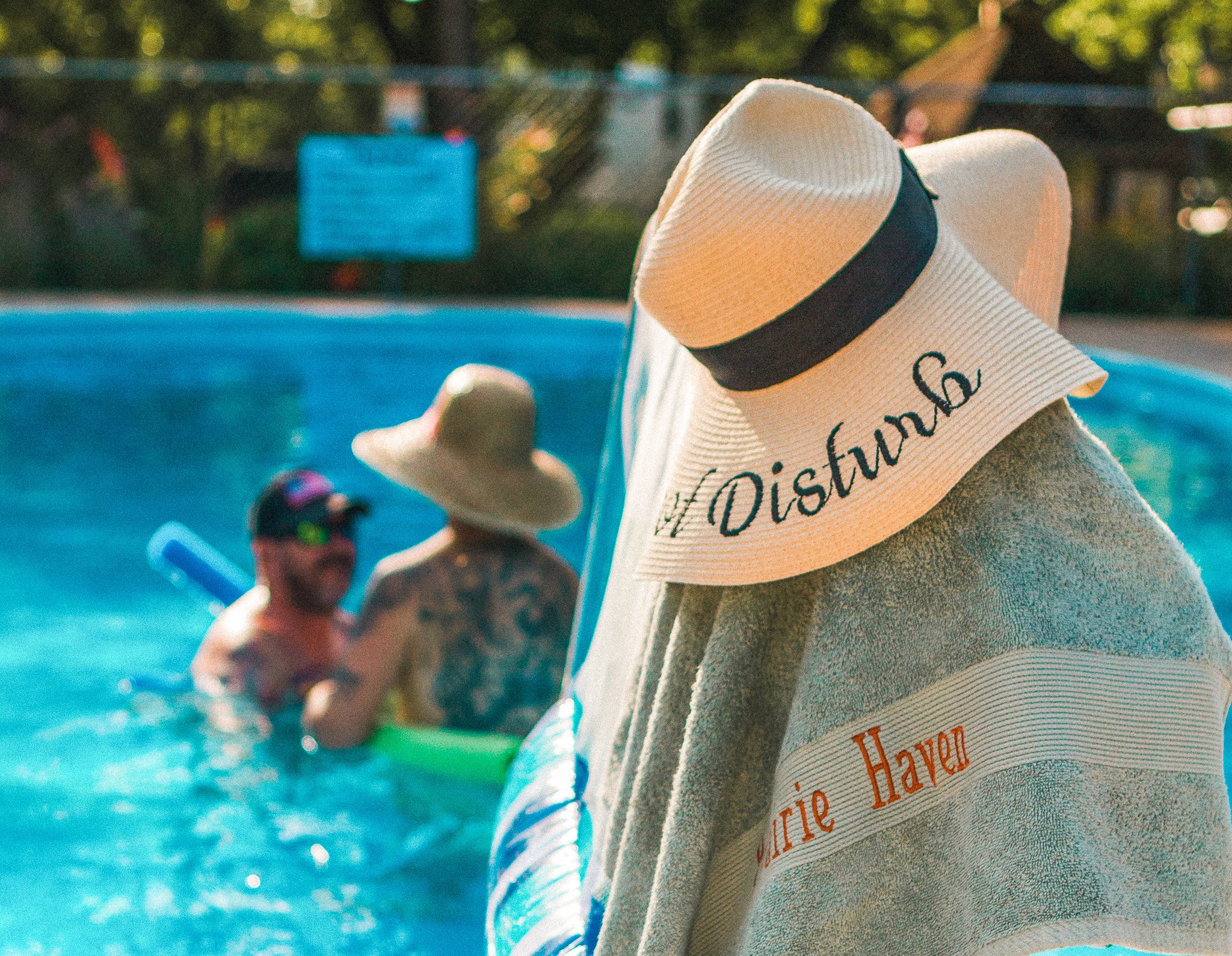 Person with a wide-brimmed hat and towel watching two people swimming in an outdoor pool.