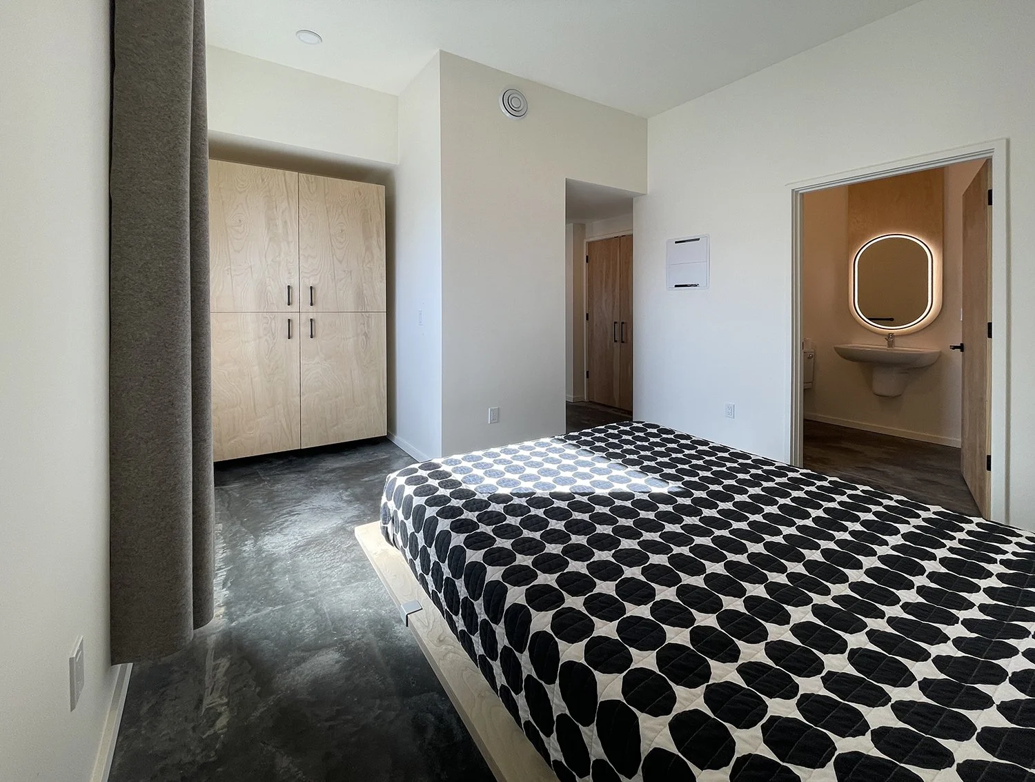 A bedroom with a bed featuring black and white polka dot bedding, a large cabinet with wooden doors, an open door revealing a bathroom with a mirror and sink, and concrete flooring.