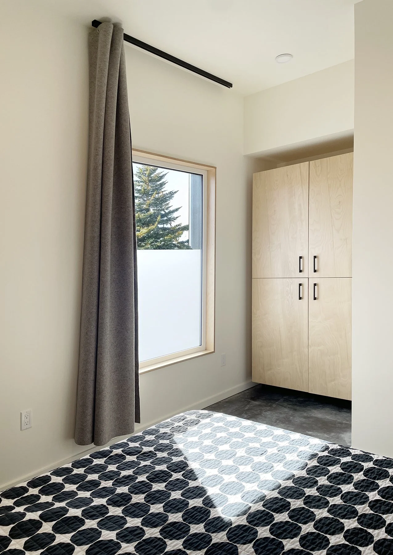 A bedroom with a bed that has a black and white patterned quilt, a window with a wooden frame and a view of trees outside, a light-colored wooden closet, a gray curtain, and a circular ceiling light.
