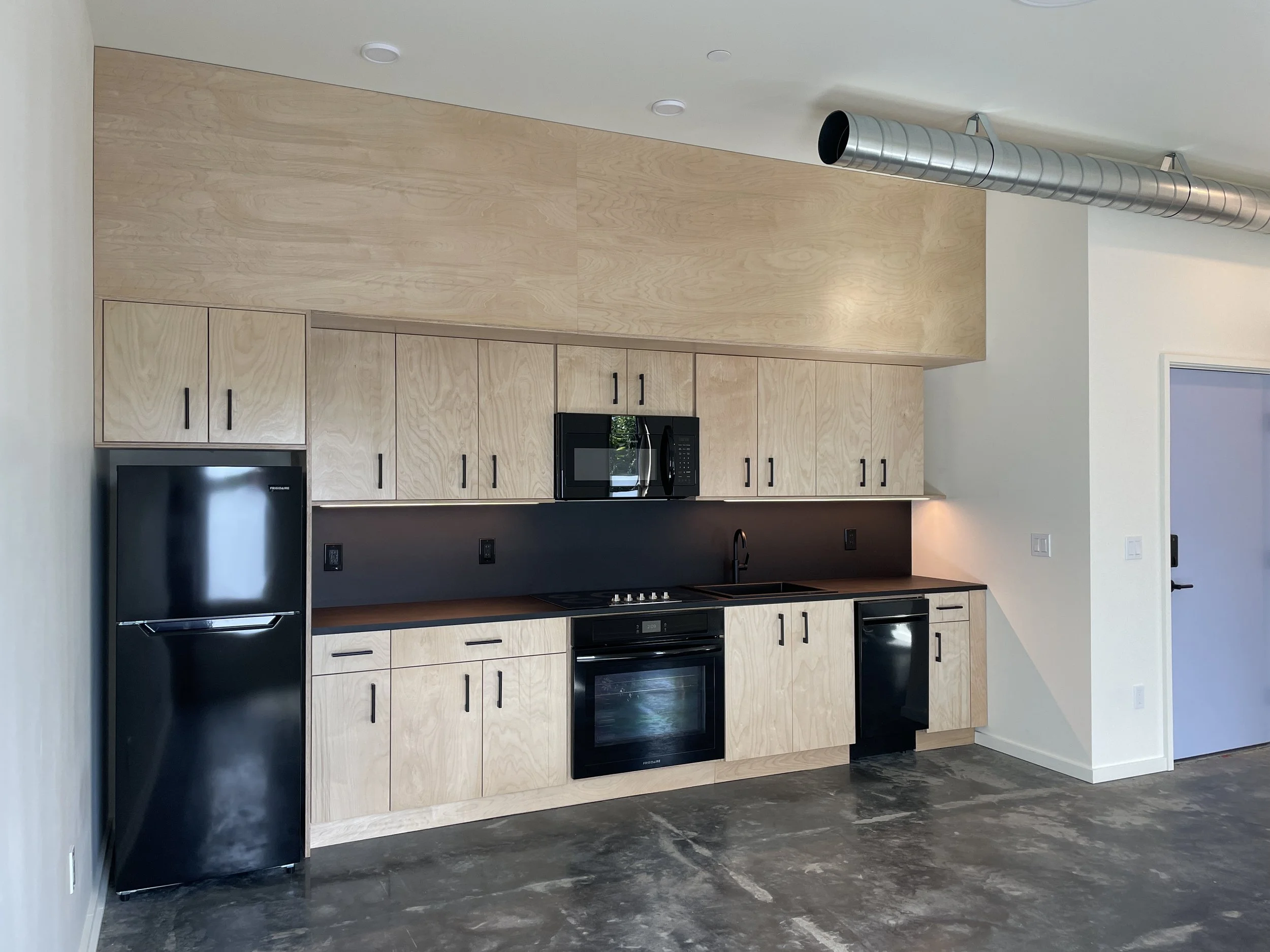 Modern kitchen with black appliances, light wood cabinets, and a black countertop. There is a large silver ventilation duct running along the ceiling.
