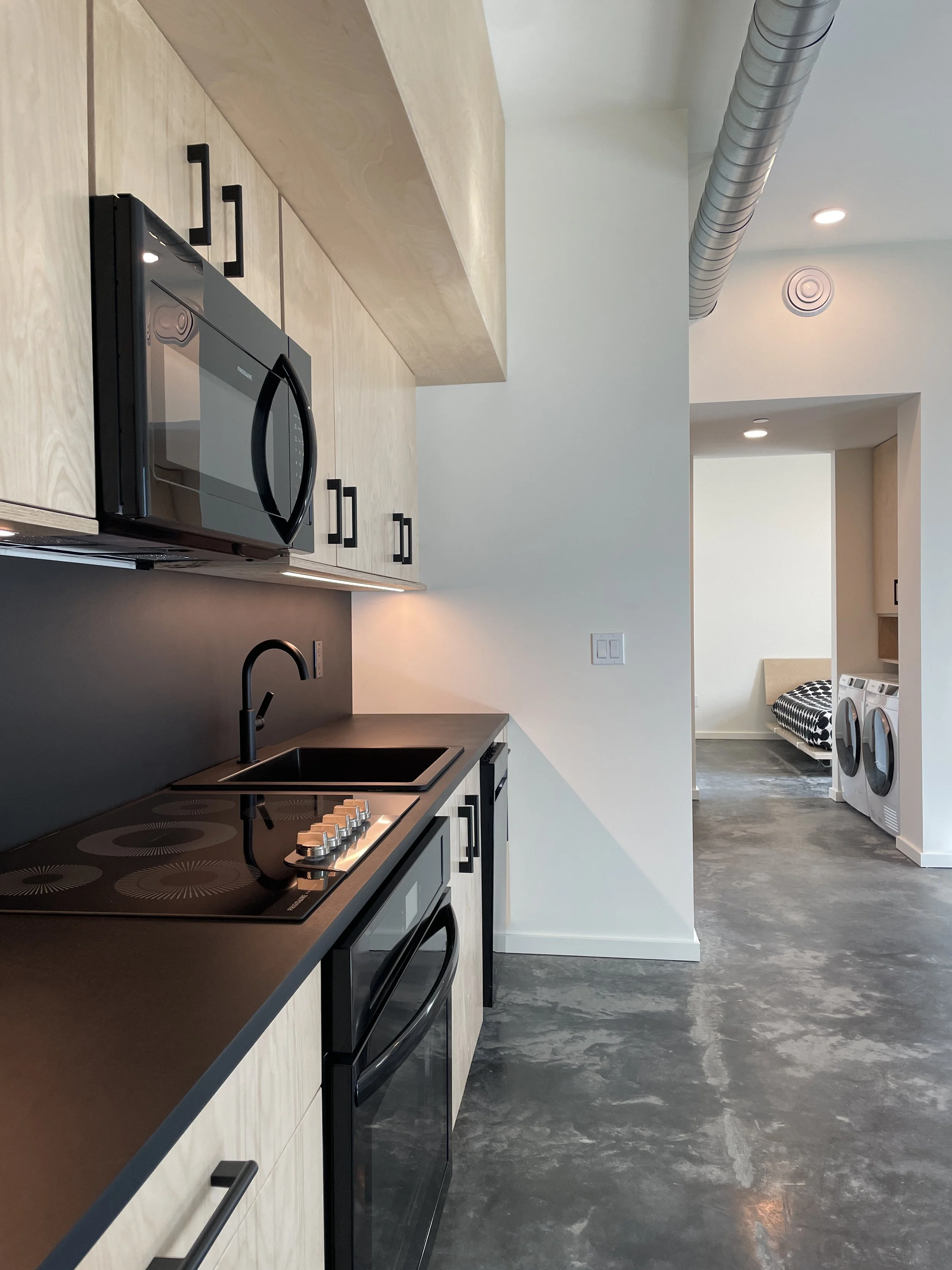 A modern kitchen with a black stove, microwave, and oven, light wood cabinets, a black faucet, and grey marble floor. In the background, there are laundry appliances and a small bed in a neighboring room.