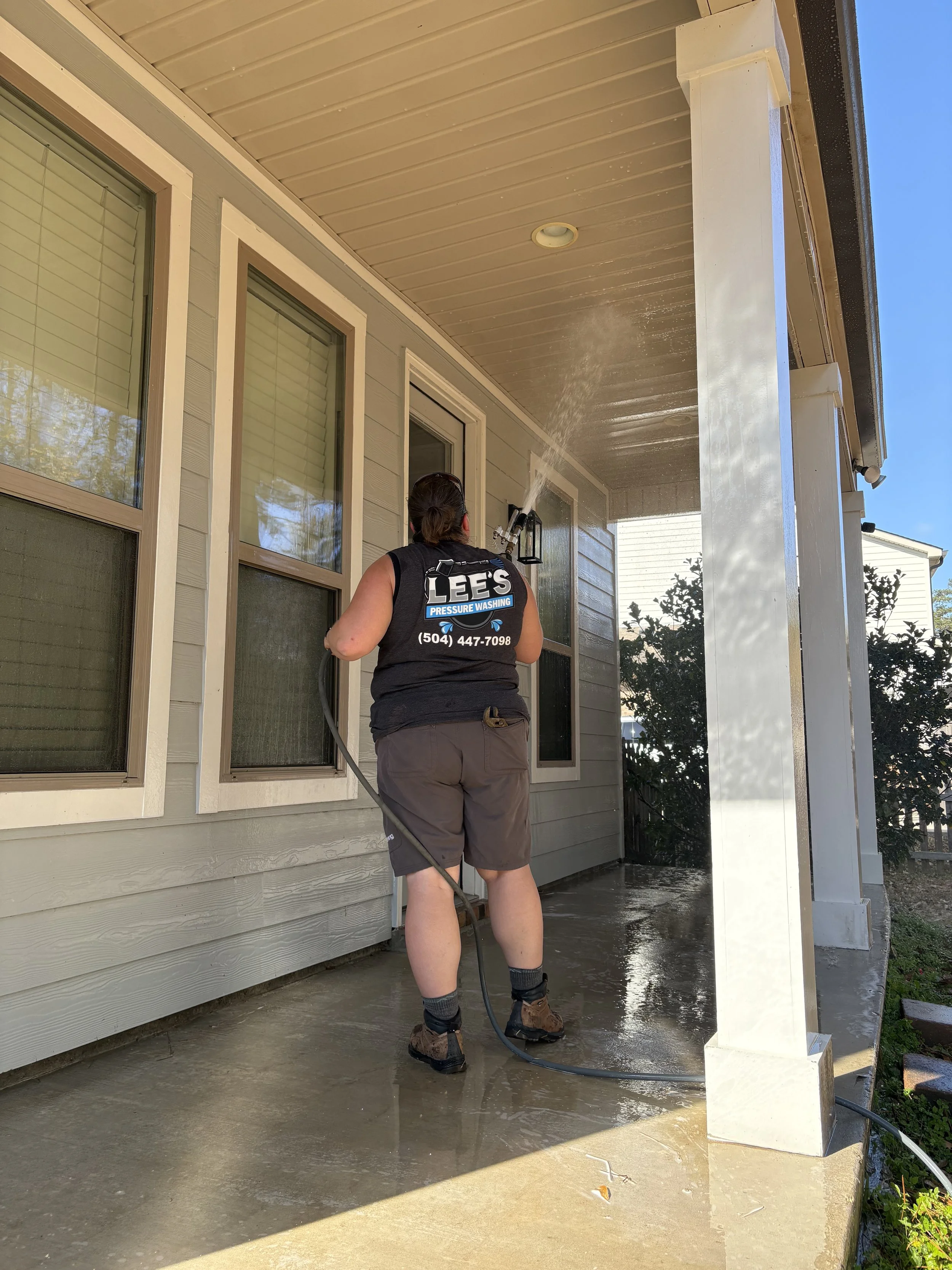A person is pressure washing the concrete porch of a house. They are wearing a black sleeveless shirt with the logo 'Lee’s Pressure Washing' on the back, gray shorts, and work boots. The house has beige siding and large windows with closed blinds. Water from the pressure washer is spraying the porch, which appears wet, with the sunlight creating reflections on it.