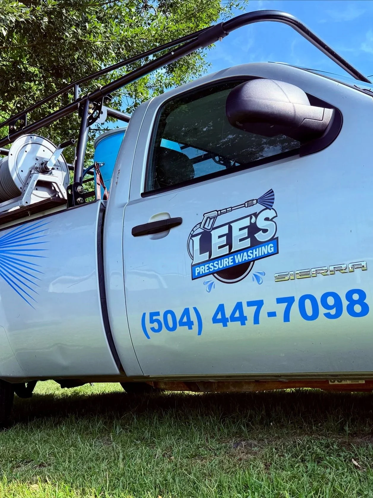 Pickup truck with company logo 'Lee's Pressure Washing' on the side, parked on grass under a tree, carrying cleaning equipment and hoses in the truck bed.
