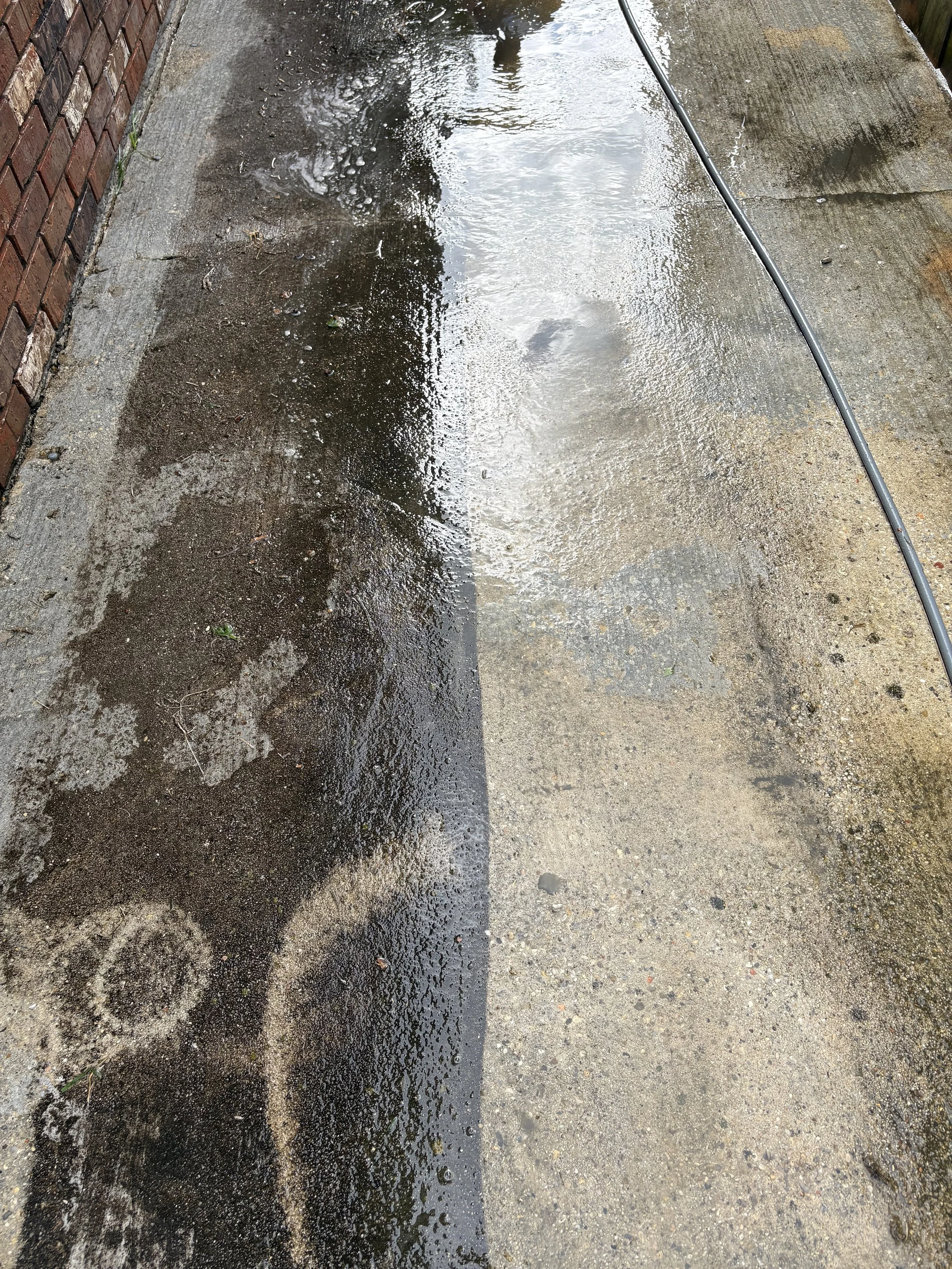 A concrete sidewalk with a dirty black patch on the left side and a lighter clean patch on the right side, next to a brick wall and a garden hose.