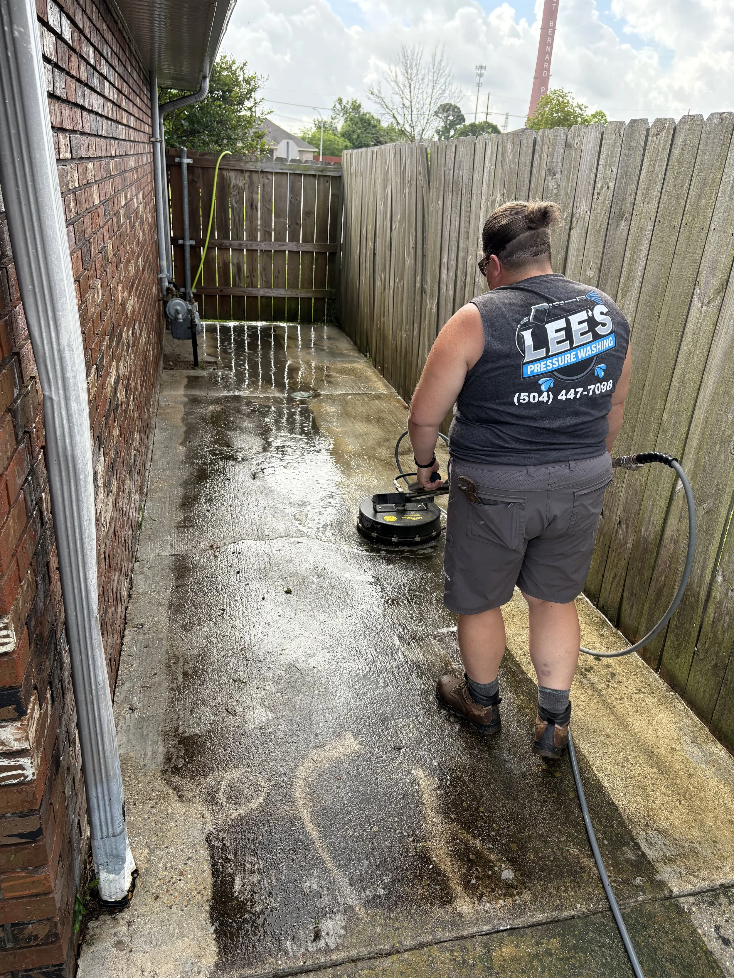 A person in work clothes pressure washing a concrete patio outside a house, with a wooden fence and brick wall visible.
