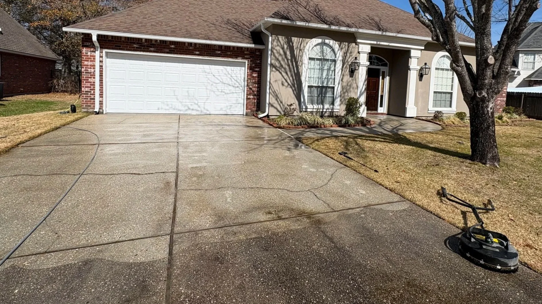 Front yard of a house with a concrete driveway, a tree on the right, a flower bed near the front door, and a white garage door. A cord and a robotic lawn mower are on the driveway.