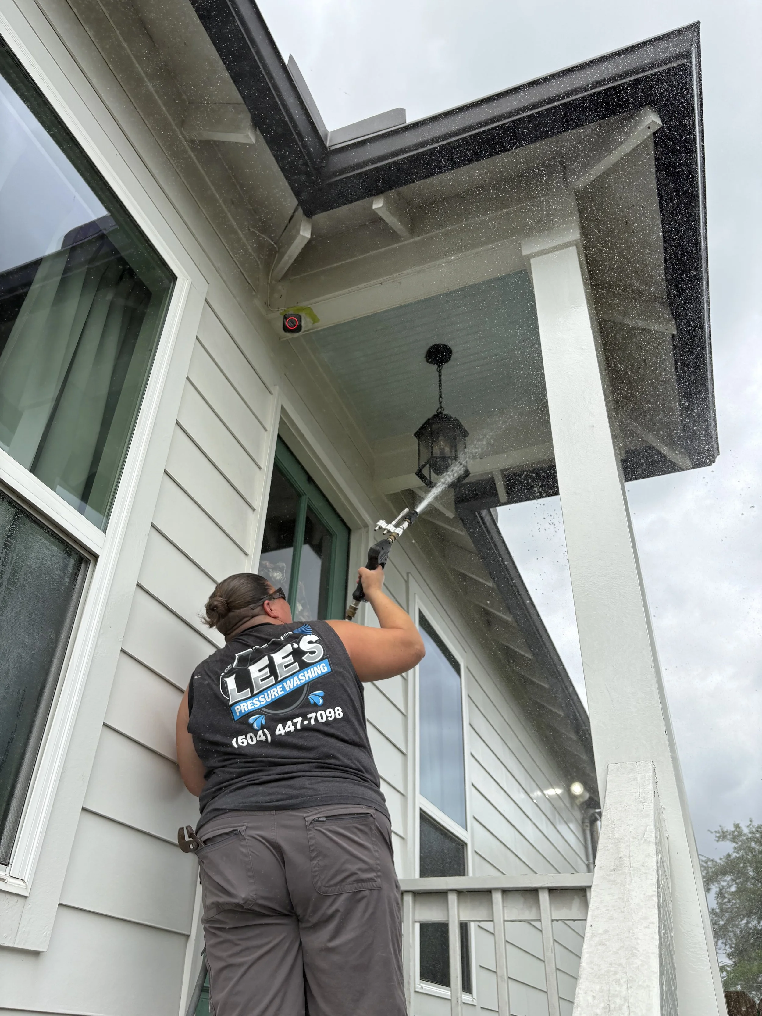 A person cleaning a house's porch from outside using a pressure washer, with a visible logo on their shirt that reads "Lee's Pressure Washing" and a phone number.