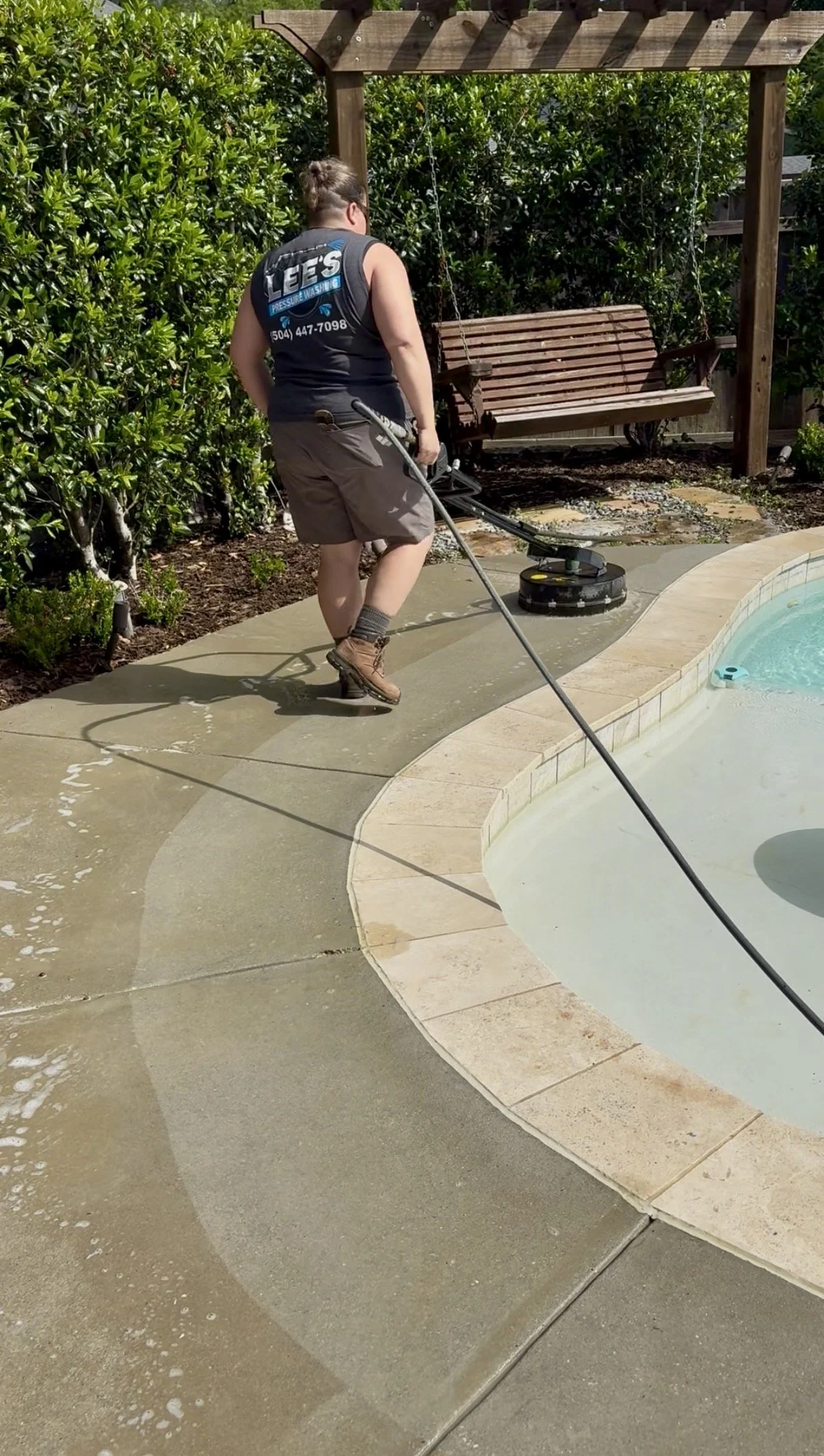 Worker using a pressure washer to clean the concrete patio around a swimming pool, with a wooden swing in the background, surrounded by bushes.