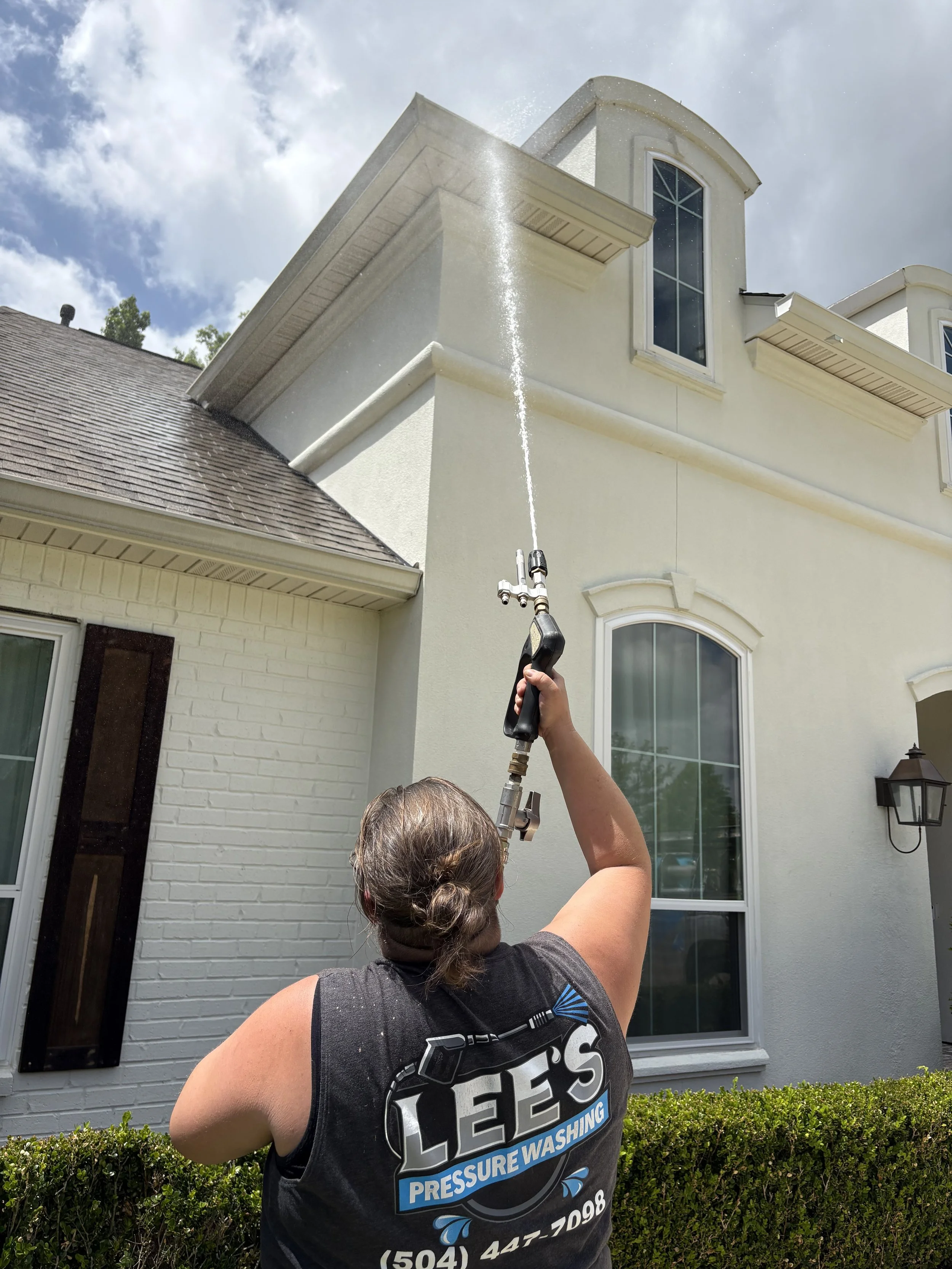 A woman with curly hair wearing a black tank top with logo cleaning the exterior window of a house with a pressure washer, with the house's white wall and large window behind her.