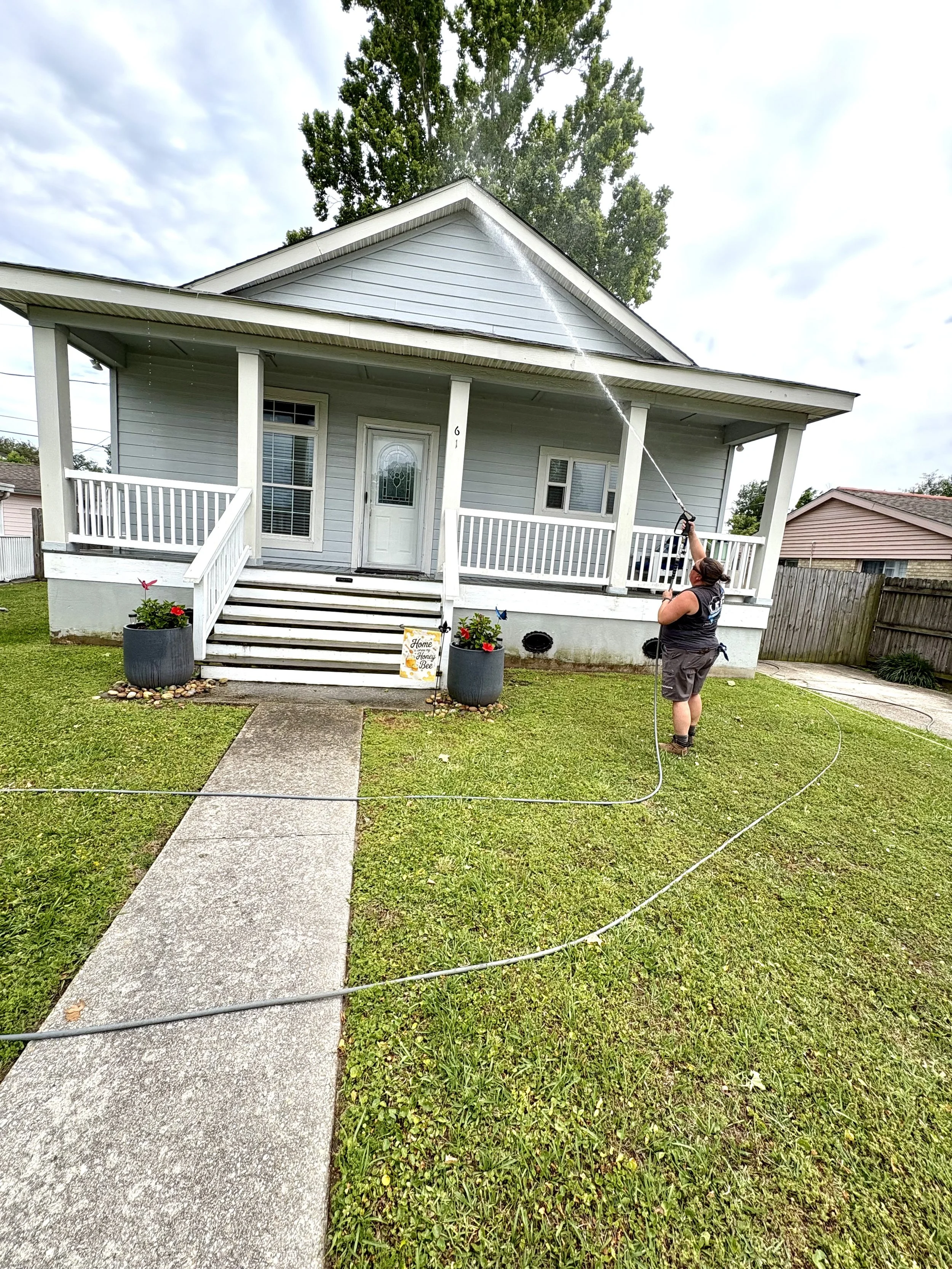 A person spraying water on the front peak of a house with a pressure washer, with a cloudy sky and neighboring houses visible in the background.