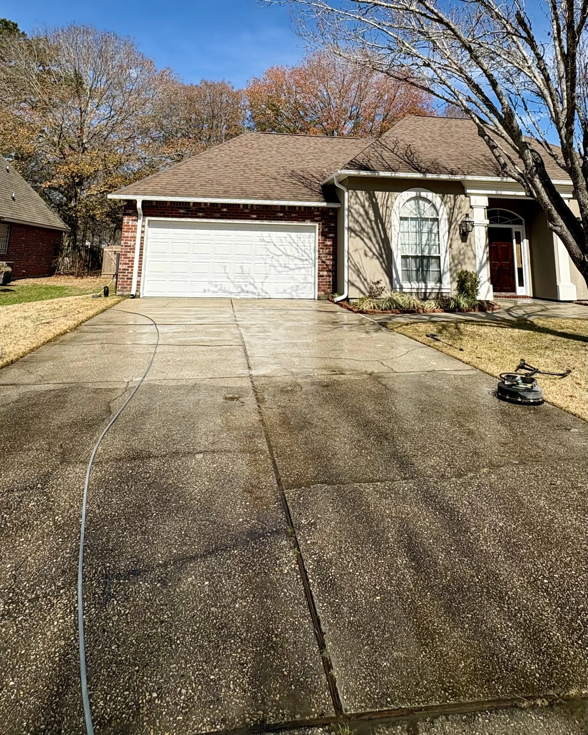Driveway with top half clean and bottom half still dirty in front of a house with a two-car garage, tree shadows on the walls, and a surface cleaner on the driveway.