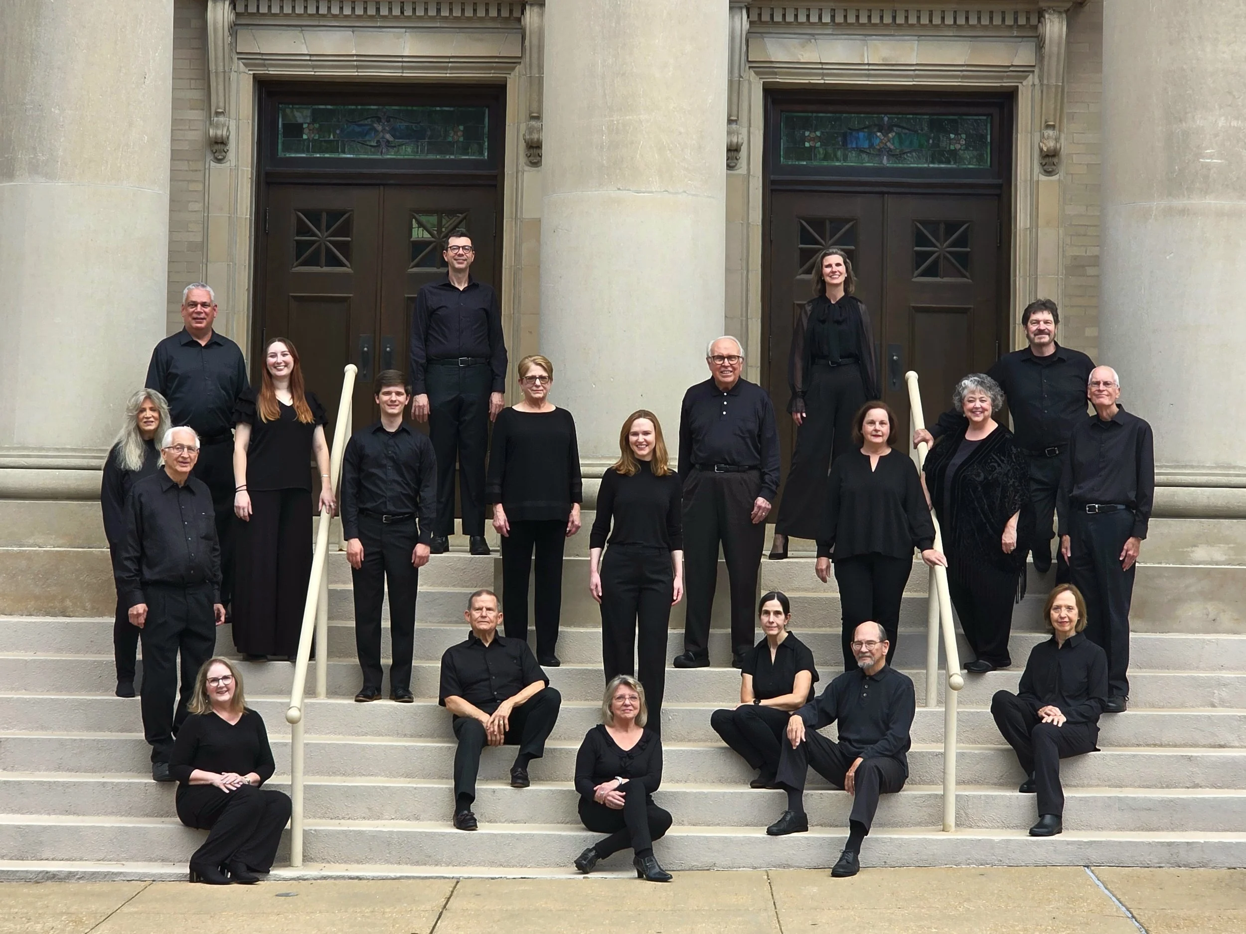 Group photo op people, likely a choir, standing and sitting on steps in front of a building with large columns and two wooden doors, dressed mostly in black.