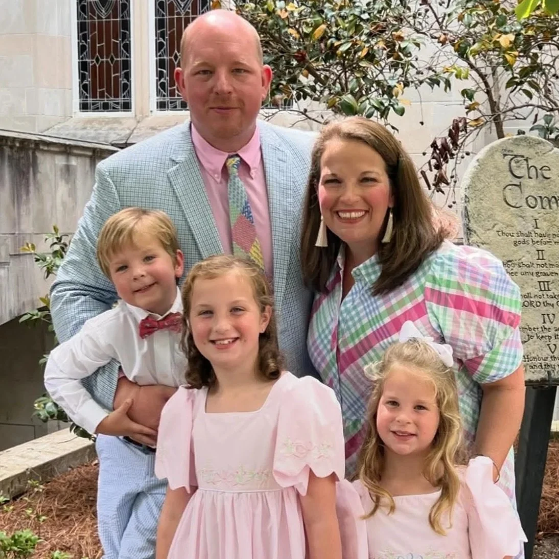 A family of five posing outdoors, with a man, woman, and three young kids, smiling in front of a stone monument and a building with stained glass windows.