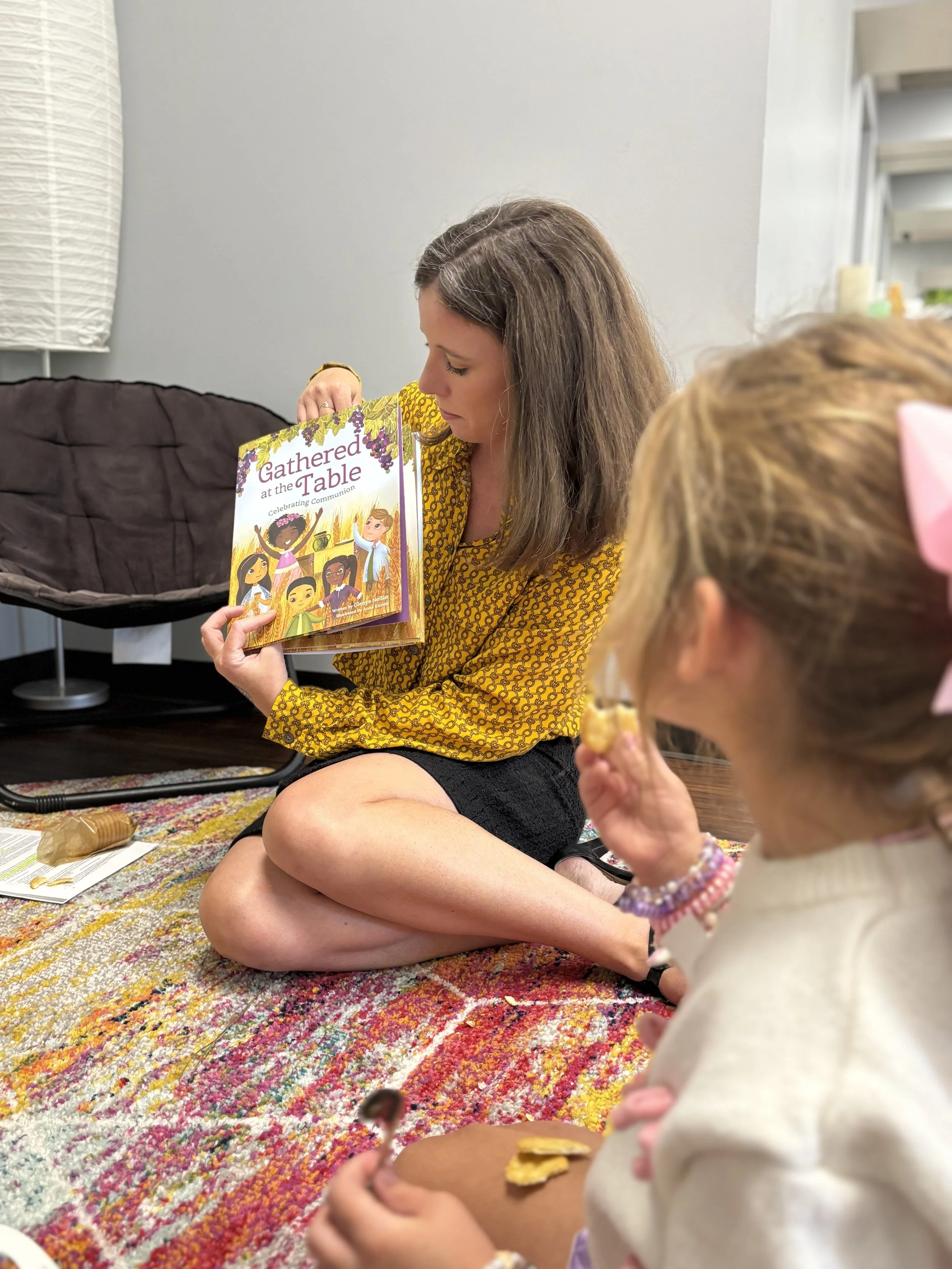 A woman sitting cross-legged on a multicolored rug, reading a children's book titled "Gathered at the Table" to a young girl wearing a cream-colored top and a pink bow in her hair, holding a snack.