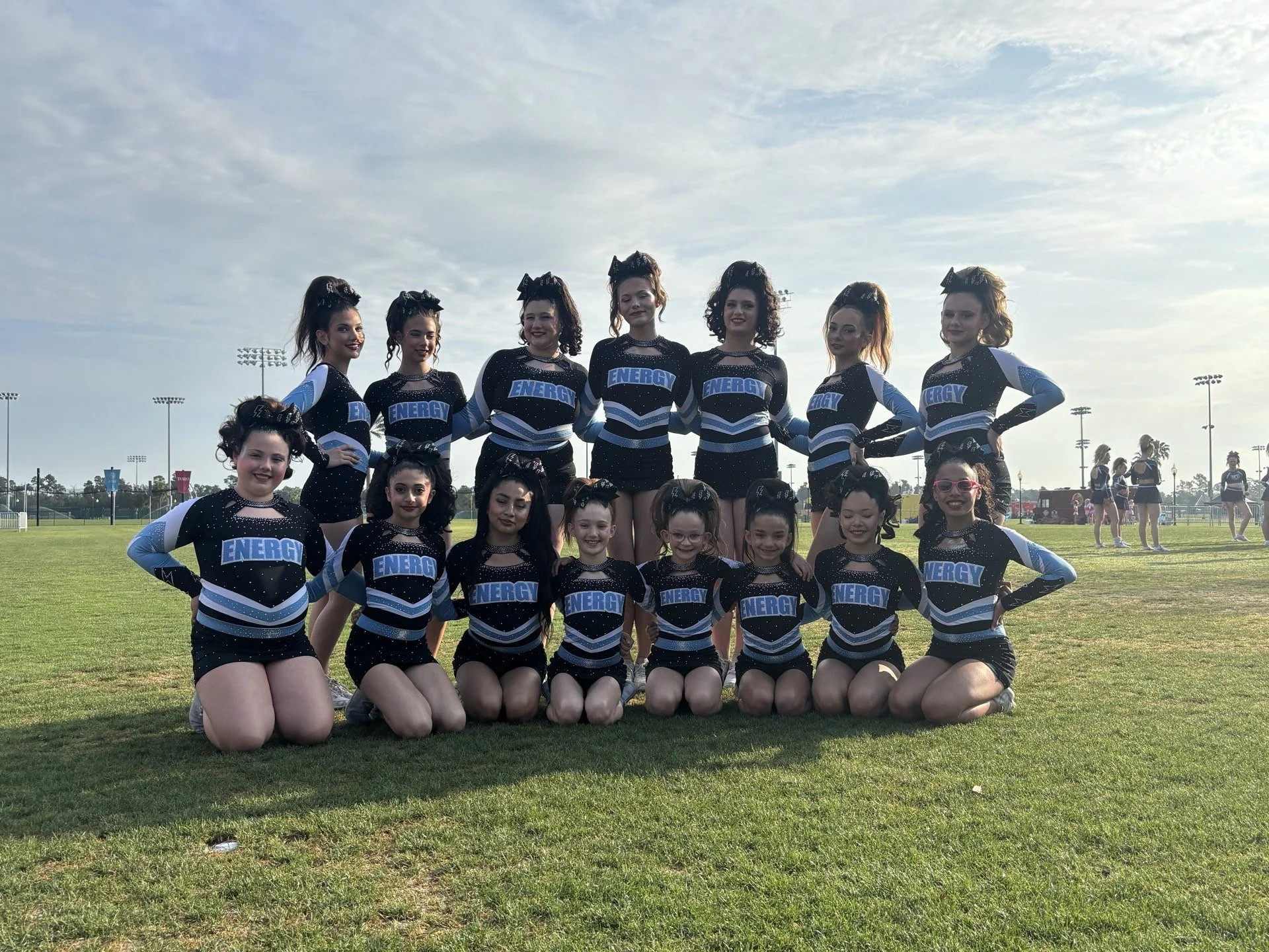 Group of cheerleaders in black and blue uniforms with 'ENERGY' written on the front, posing outdoors on a field with a cloudy sky and stadium lights in the background.