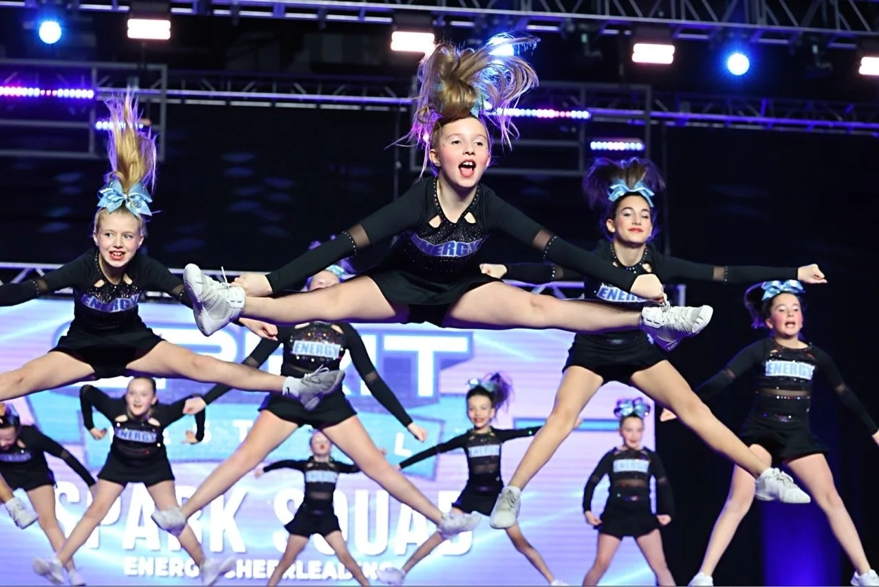 Cheerleaders performing a jump routine on stage with purple and blue lighting.
