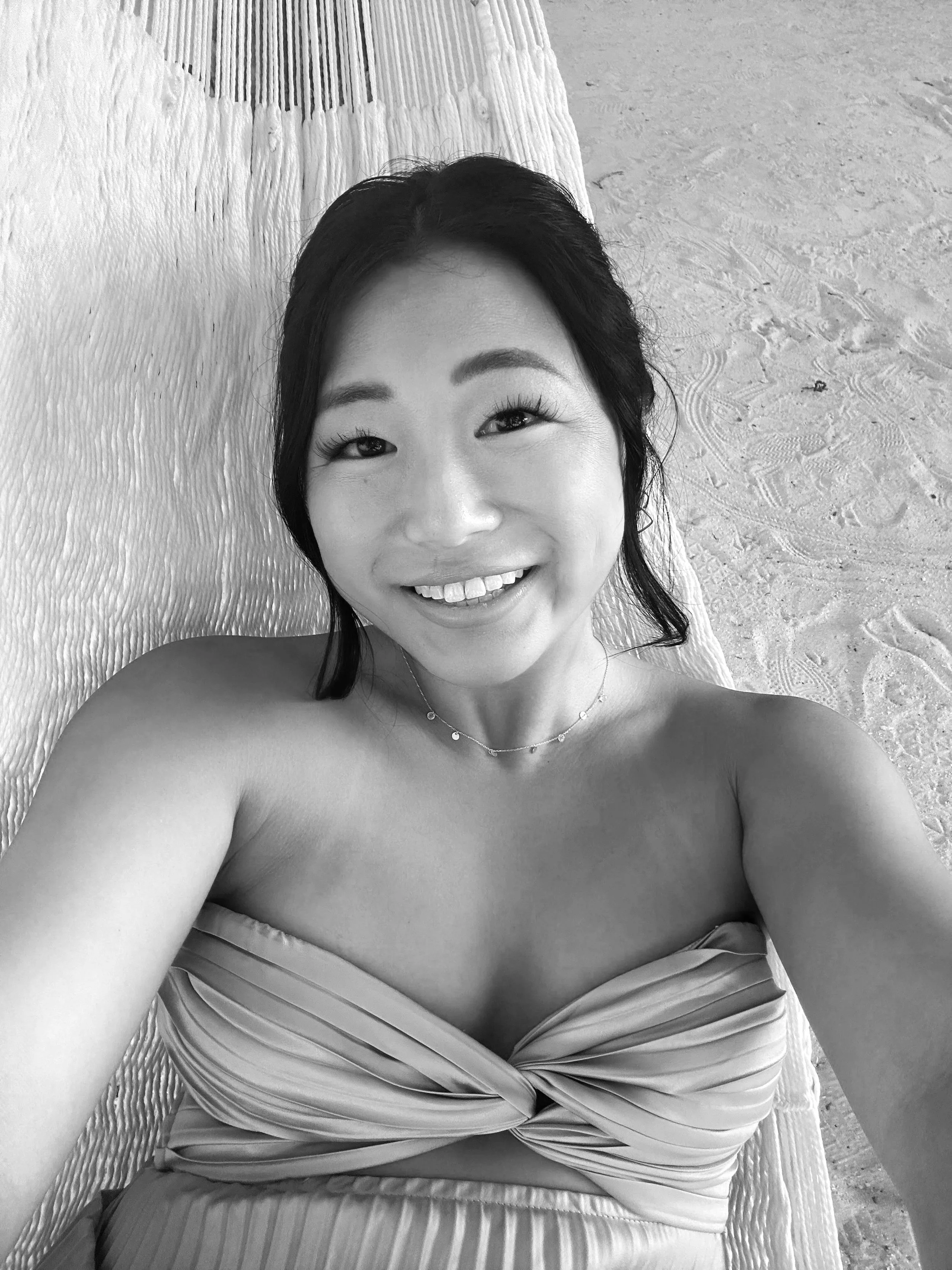 A black and white photo of a smiling woman with dark hair, wearing a strapless top and a necklace, lying on a textured surface that looks like a beach towel or mat, with sand visible in the background.