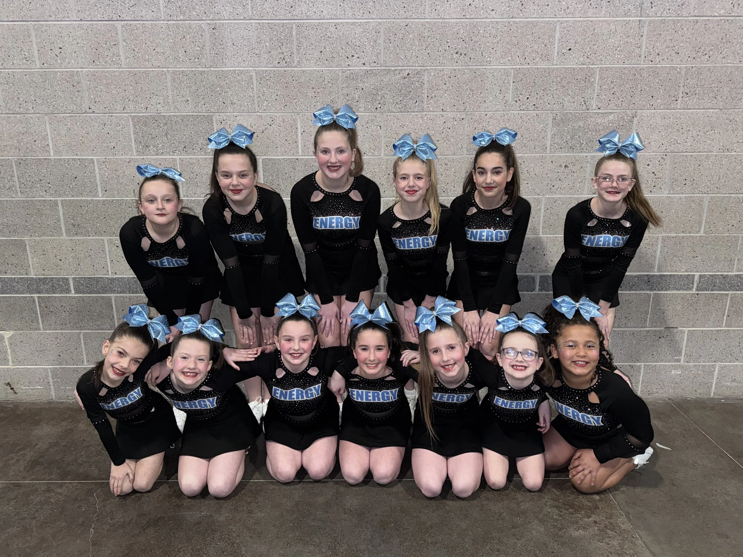 Group of young cheerleaders posing together against a brick wall, wearing black uniforms with blue bows and the word 'ENERGY' on their tops.