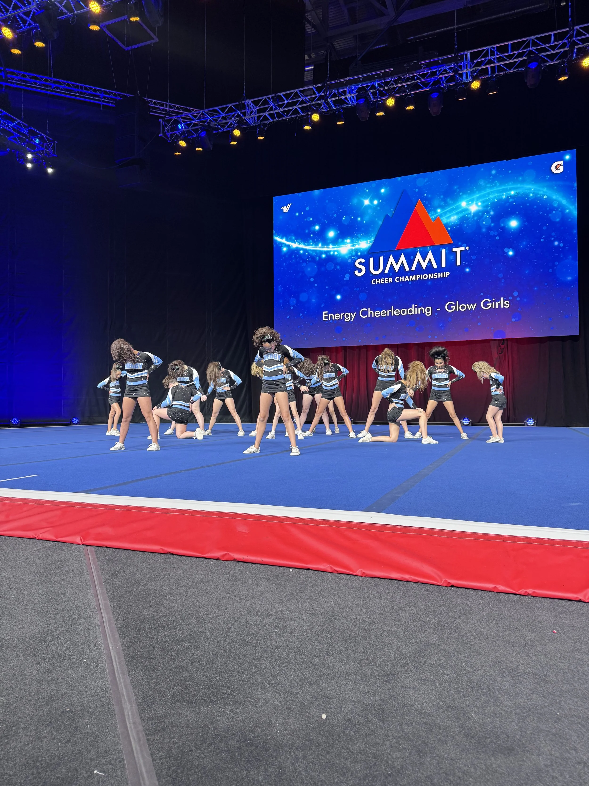 A group of cheerleaders in blue, black, and white uniforms performing a routine on a blue mat at a cheerleading competition. The large screen behind them displays the event name 'SUMMIT Cheer Championship' and the categories 'Energy Cheering - Glow Girls'.