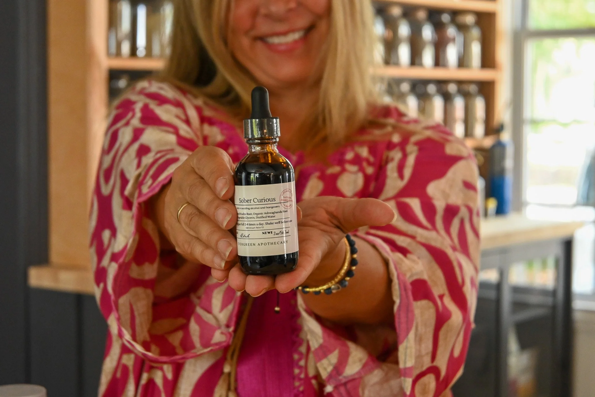 A smiling staff member extends a small amber bottle labeled “Sober Curious” toward the camera, showcasing one of Apotëak’s herbal supplement tinctures.