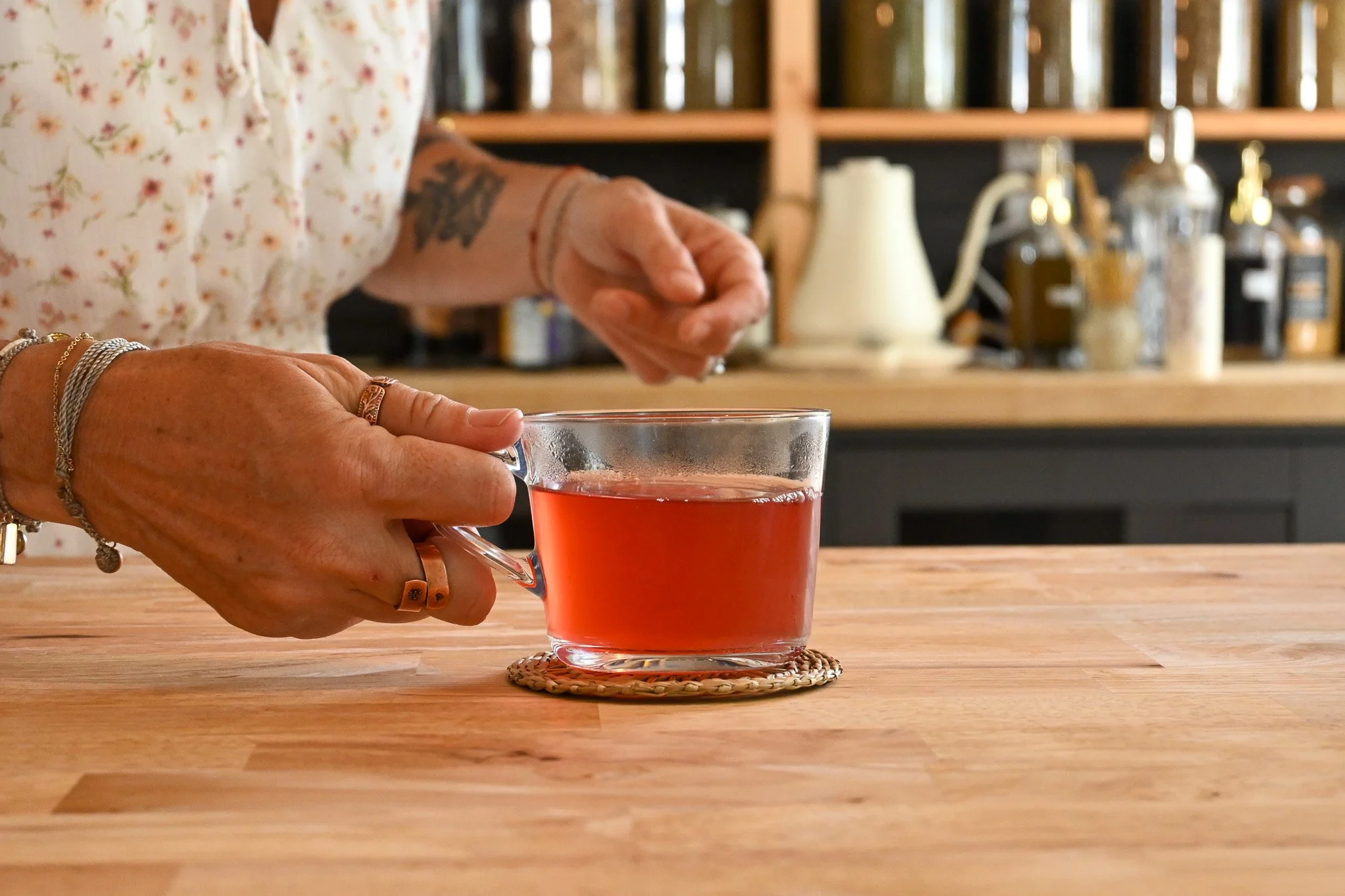 A pair of hands gently sets down a clear glass teacup brimming with deep red herbal tea on a wooden counter, representing Apotëak’s freshly brewed tea blends.