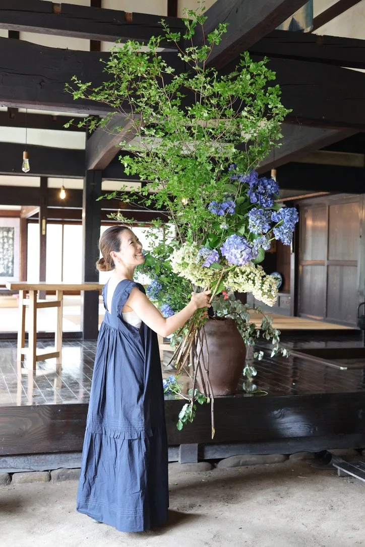 Woman in a blue dress holding a large bouquet of blue and white hydrangeas indoors.