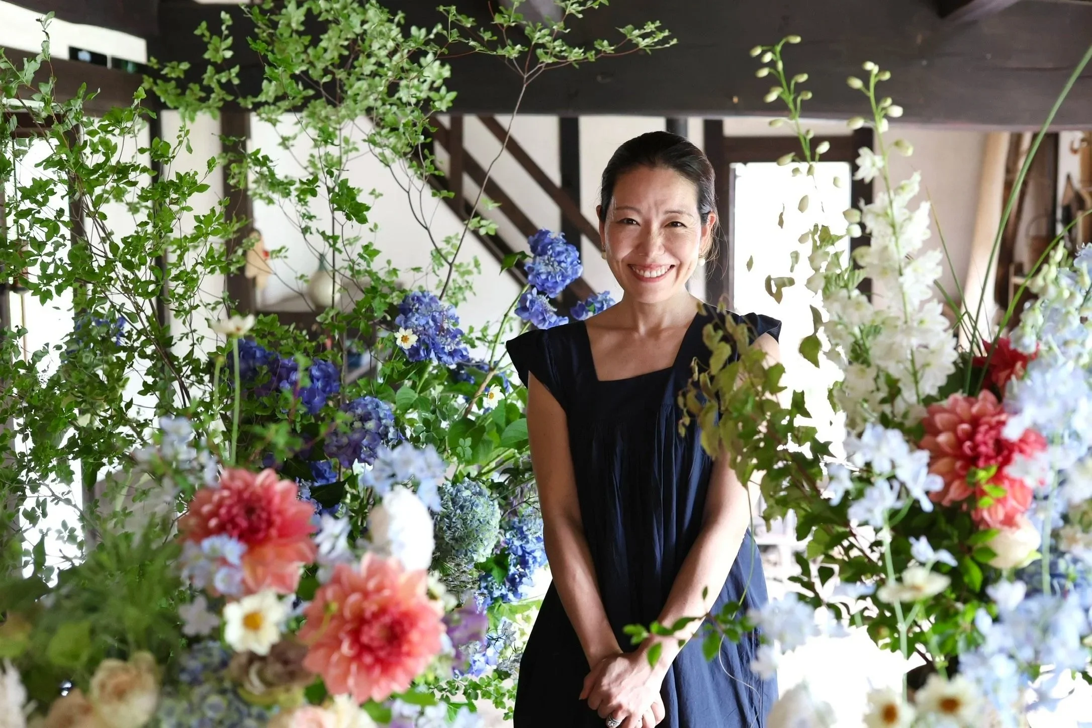 A woman in a black dress smiling, surrounded by colorful flowers and green foliage inside a room with a staircase in the background.