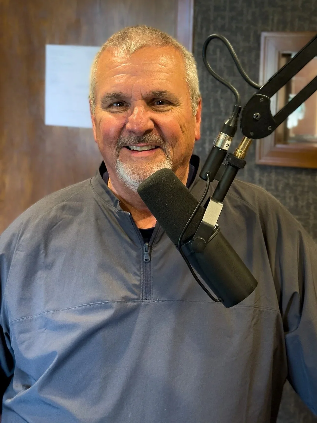 A man with gray hair and a beard, wearing glasses, a plaid shirt, and a dark zip-up sweater, standing indoors with a wooden wall and radio station signage in the background.