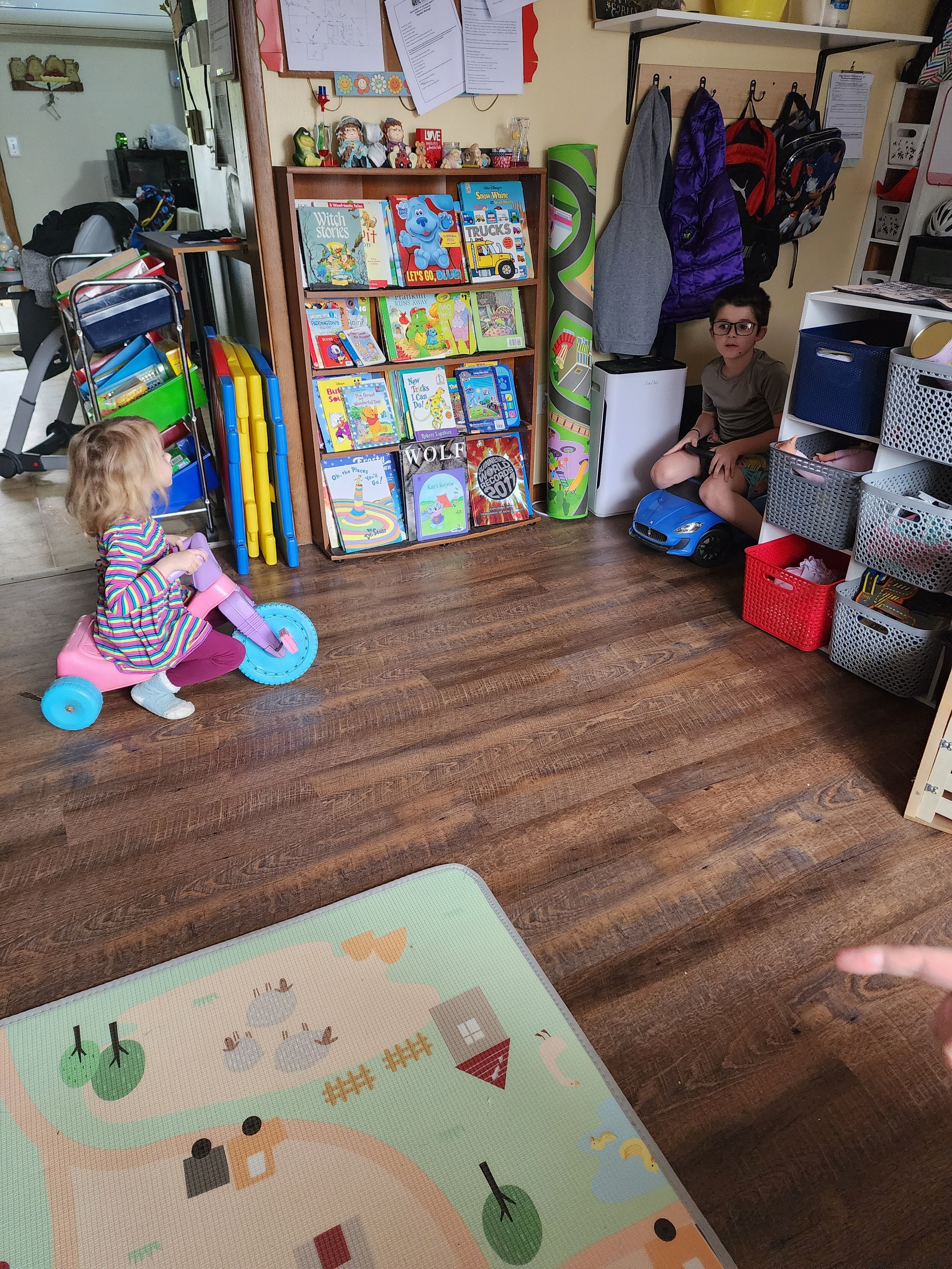 A room with two children playing. One child rides a pink tricycle while the other sits on a blue toy car. Nearby is a shelf filled with children's books and toys. Coats and backpacks hang on hooks, and a colorful play mat is on the wooden floor.