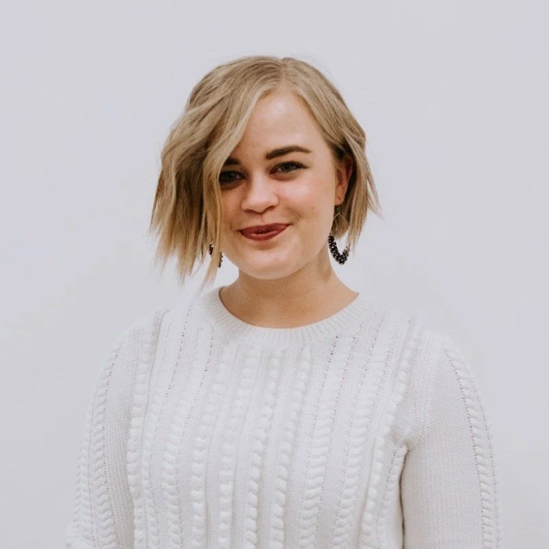 A woman with blonde, messy hair wearing a white cable-knit sweater and dark earrings, smiling at the camera against a plain white background.