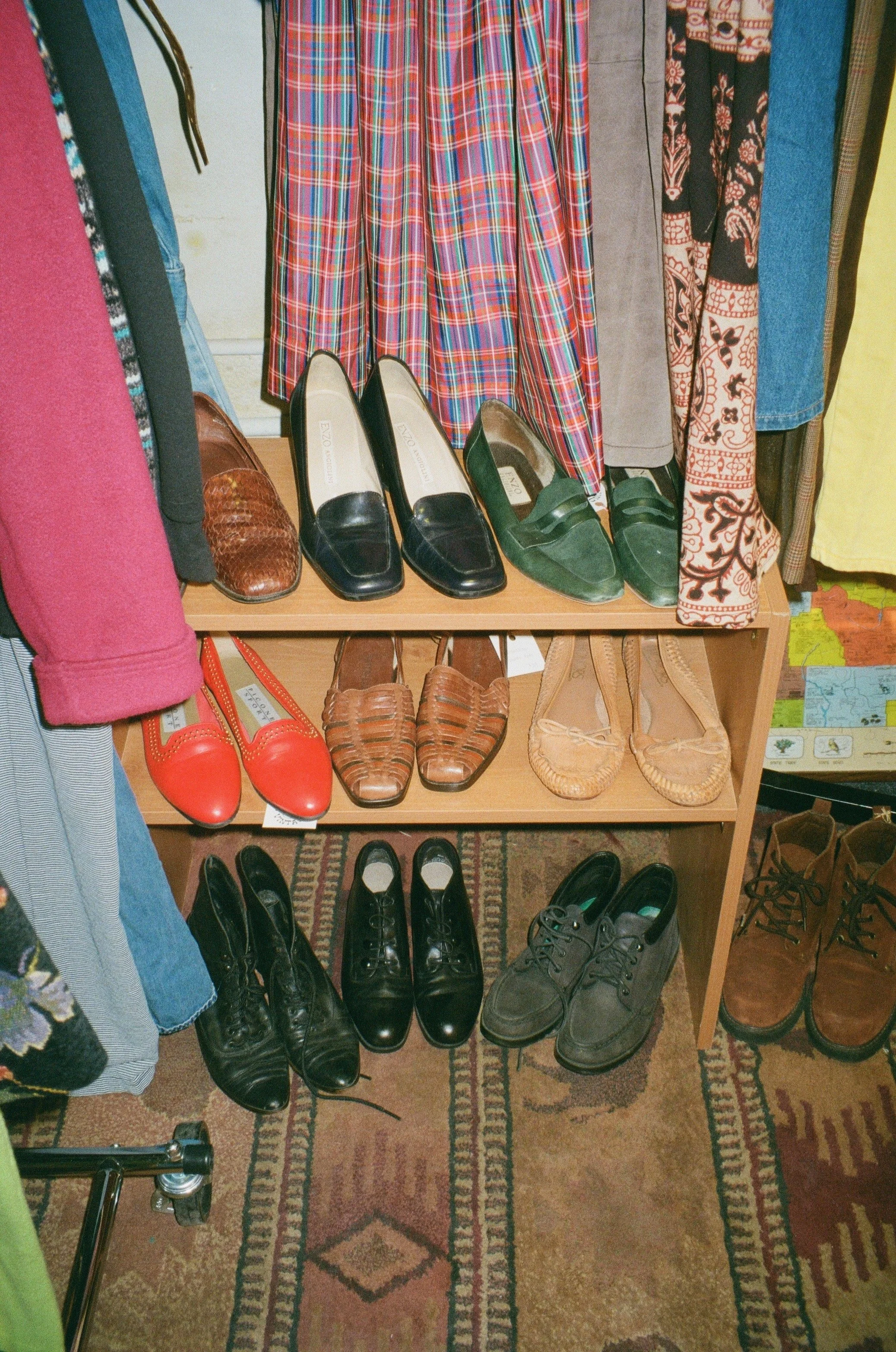 A display of various shoes on a wooden shelf with colorful clothing hanging above and to the side.