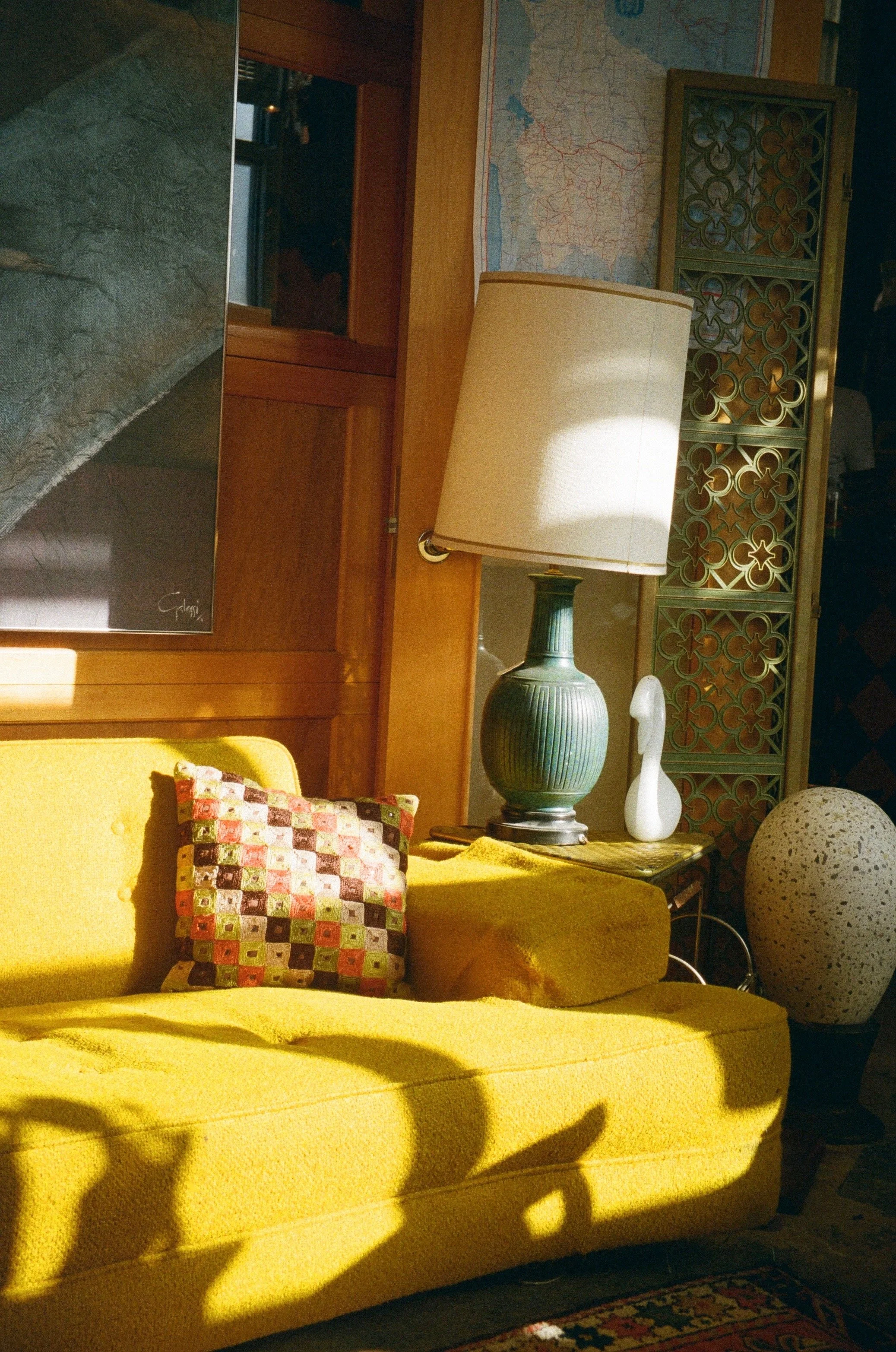 A yellow upholstered sofa with a colorful patchwork pillow, next to a dark table with a green ceramic lamp, a white decorative object, and a large speckled vase. A wooden screen, a map, and artwork are in the background. Sunlight casts shadows on the