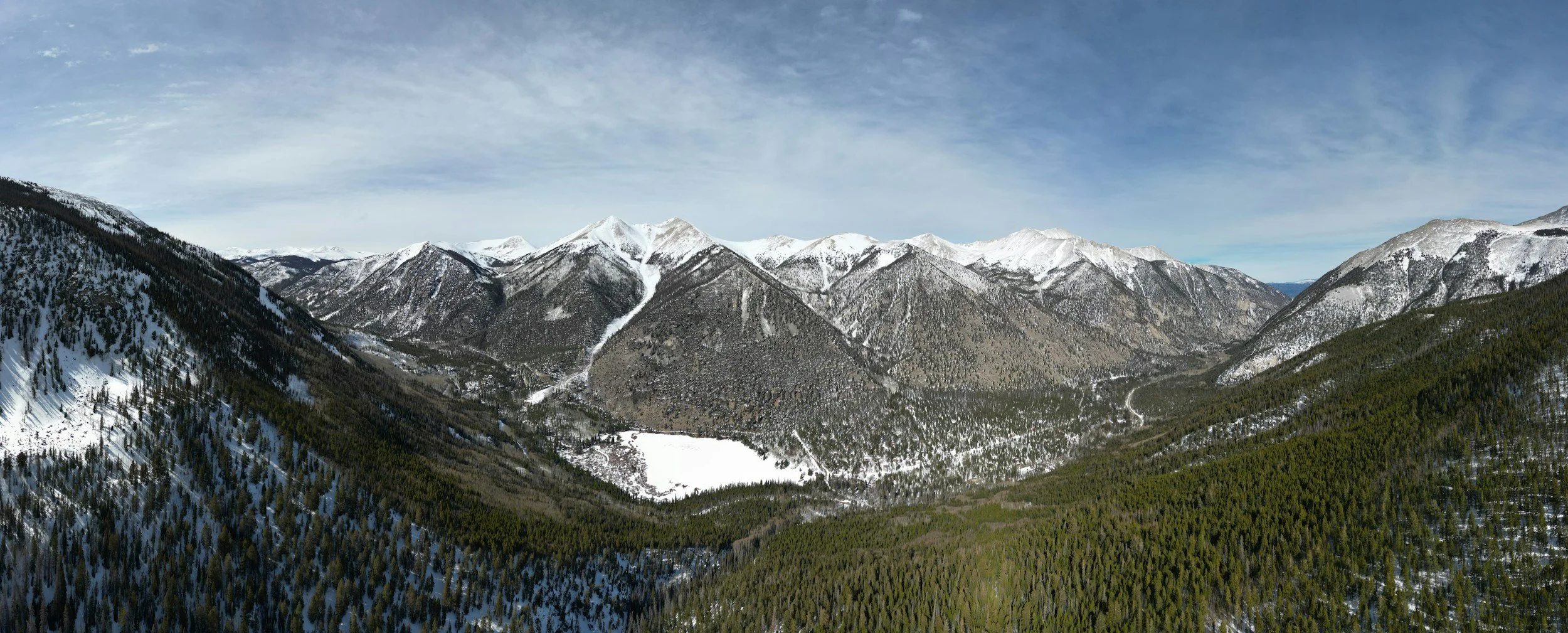 Snow-capped mountains with dense green forests in the foreground and a partly cloudy sky.