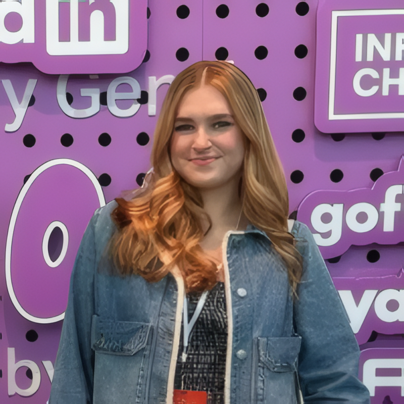 Young woman with long, wavy red hair standing in front of a purple wall with social media signs, wearing a denim jacket.