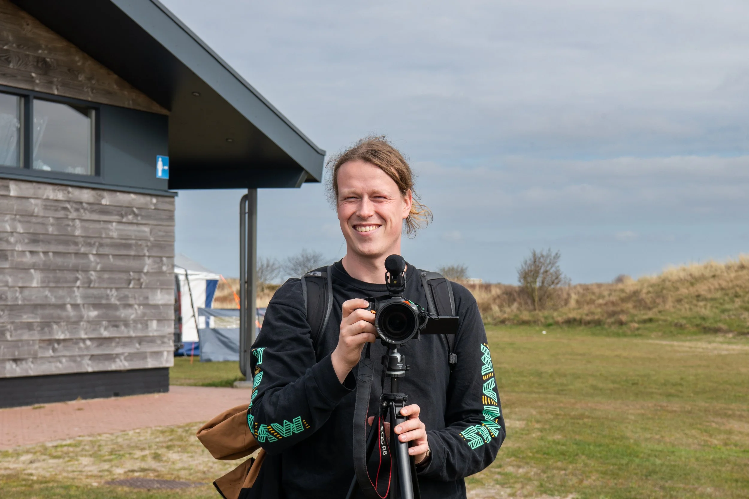 A smiling man with long hair standing outdoors in front of a wooden building, holding a camera on a tripod, wearing a black jacket with colorful text and a backpack, with a cloudy sky and grassy field in the background.
