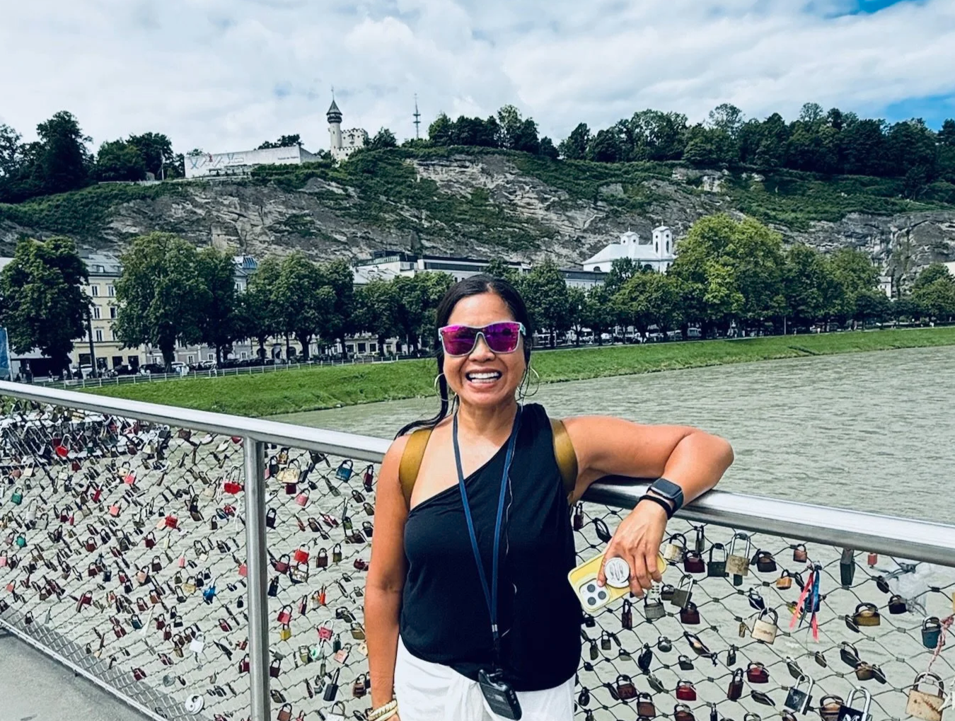 A woman smiling and standing on a bridge with love locks, holding a phone, with a river, trees, and a hill with buildings in the background.