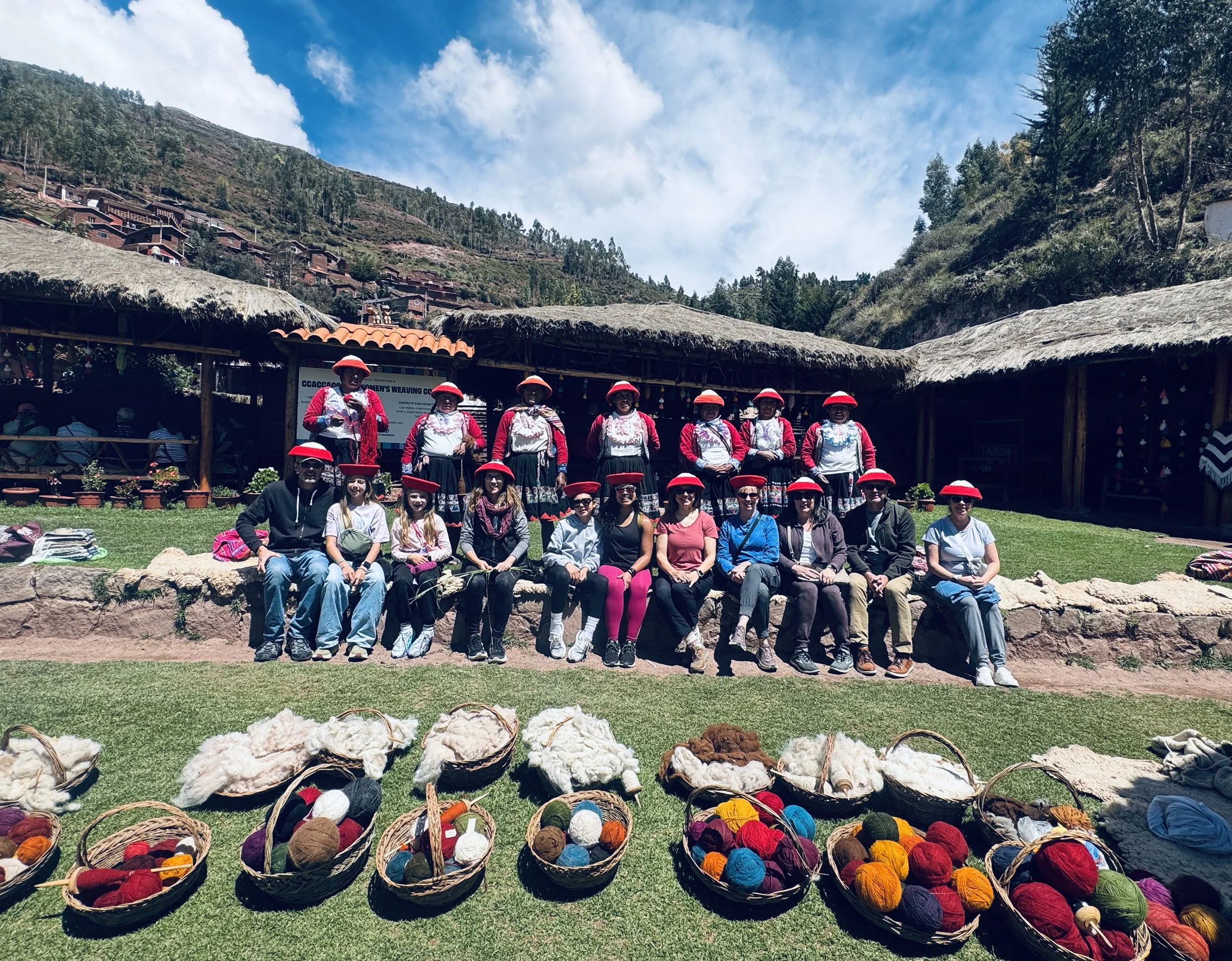 Group of people in traditional Andean clothing sitting and standing outdoors with woven baskets of colorful yarn in front, against a backdrop of thatched-roof buildings and mountainous landscape.