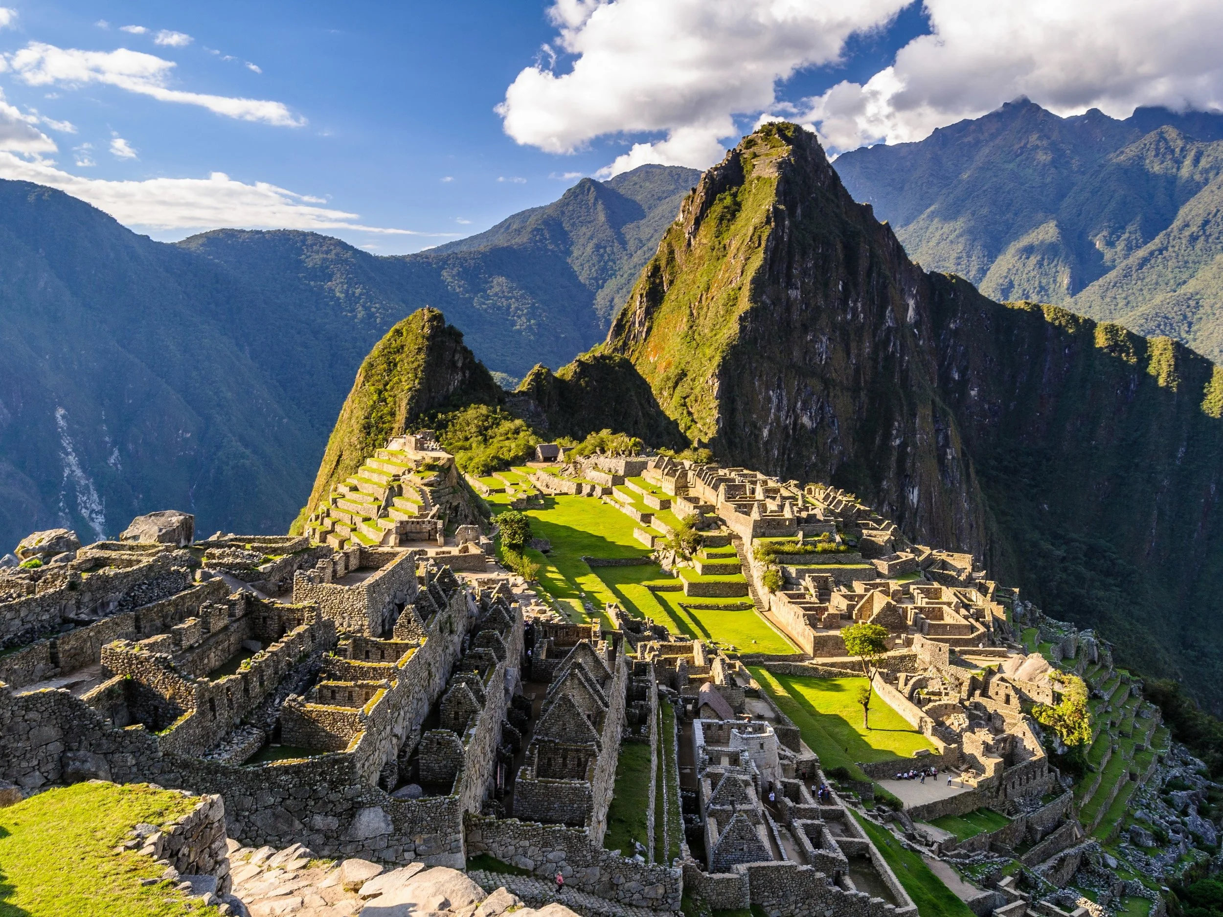 Ancient stone ruins of Machu Picchu with terraces on a mountain in Peru, surrounded by green peaks and blue sky with some clouds.