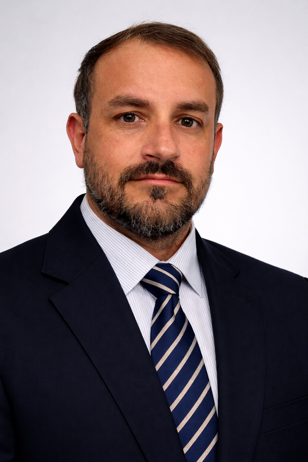 Retrato de un hombre con barba, cabello castaño y ojos marrones, vestido con traje oscuro, camisa blanca y corbata a rayas, contra fondo blanco.