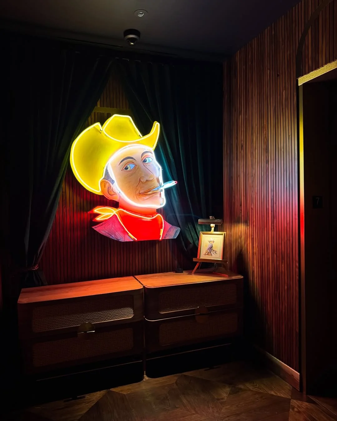 Neon sign of a cowboy's face with a cigarette in his mouth, set against a dark wood-paneled wall with curtains on either side, and a wooden cabinet below with a framed picture and small lamp on top.