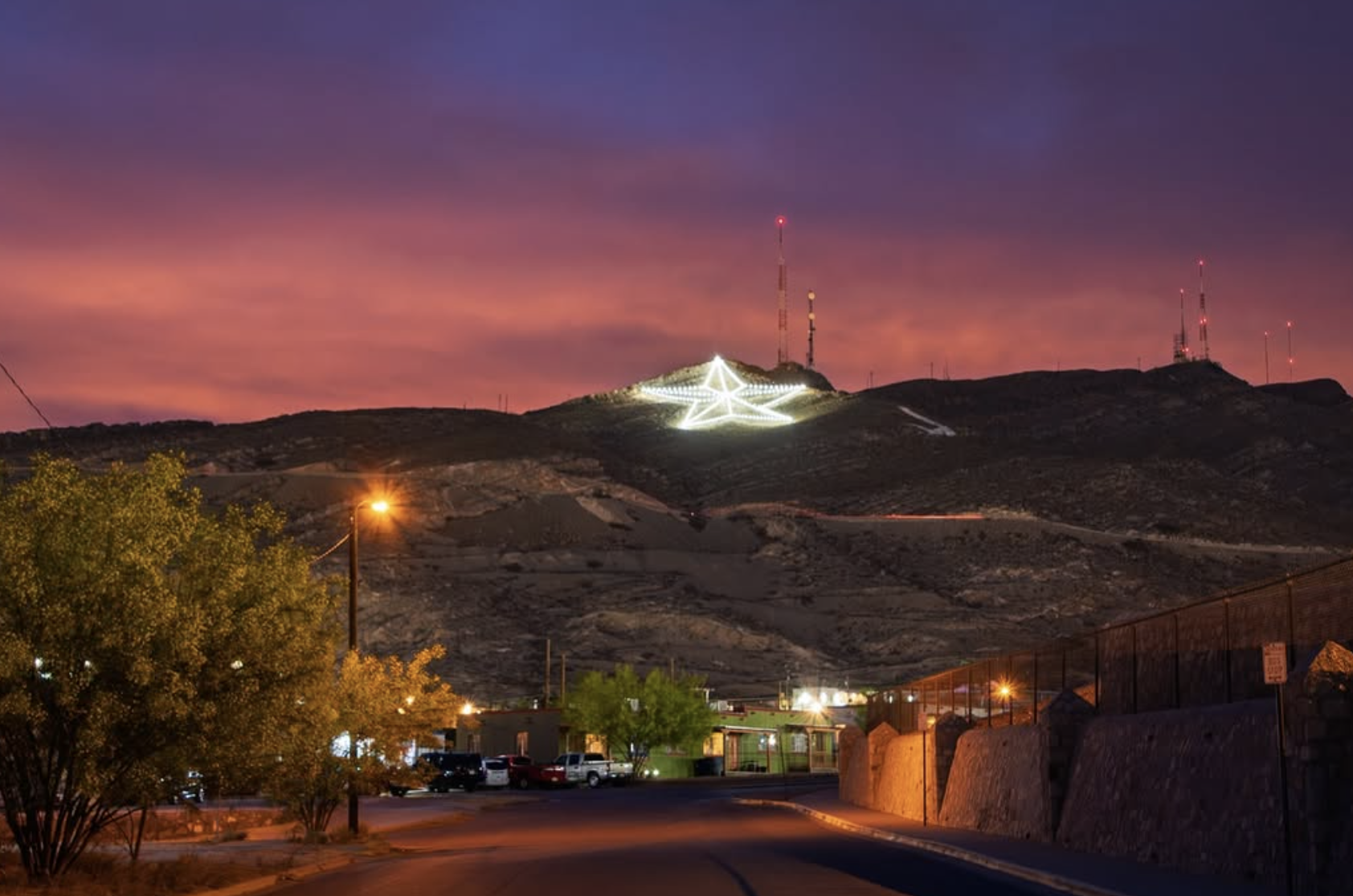 Scene of a hill at dusk with a large illuminated star decoration and red lights on tall radio towers, colorful pink and purple sunset sky, and some trees and buildings in the foreground.