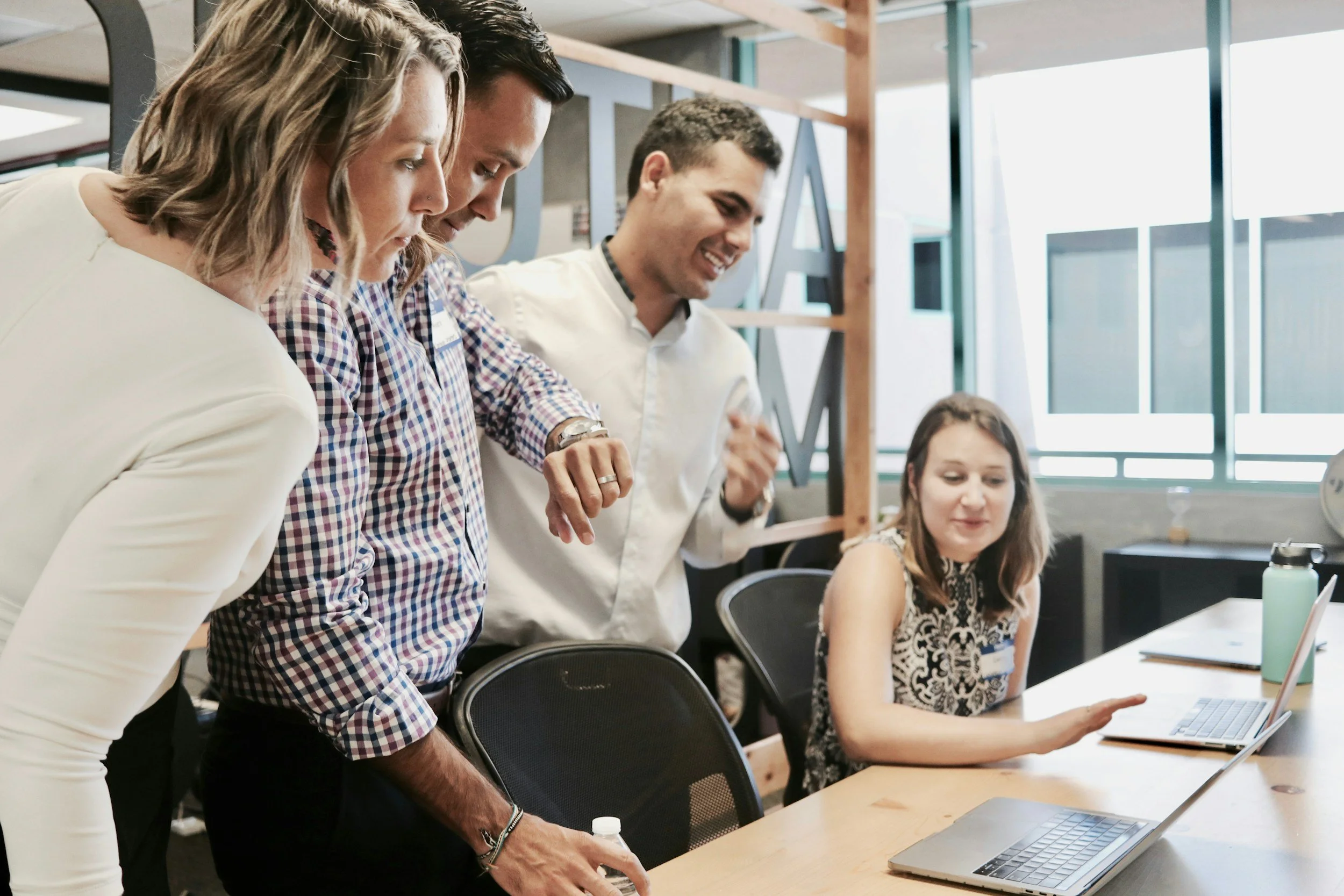 Four people gathered around a table in a modern office, looking at a laptop screen. Two men are standing, one woman is leaning over the table, and a young woman sits at the table, smiling and pointing at her laptop. The office has large windows and modern decor.