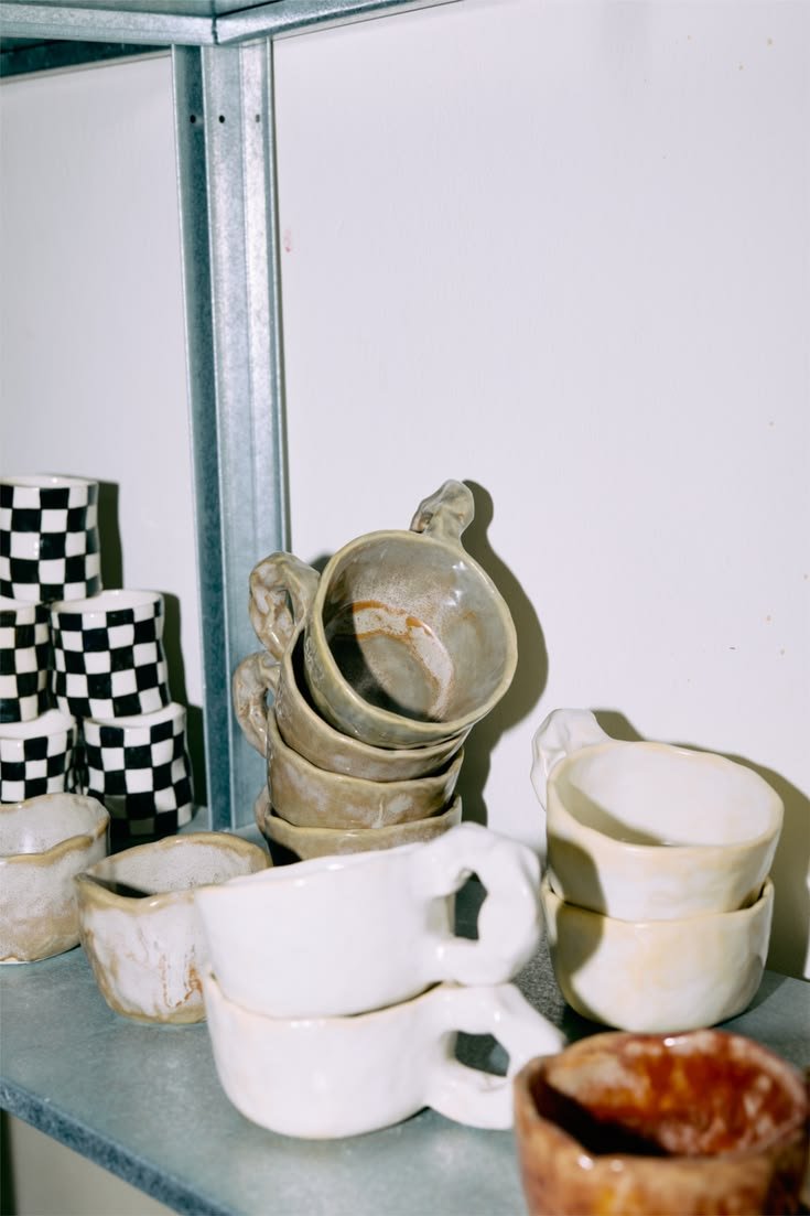 Stacked ceramic mugs and bowls on a metal shelf with a checkered box in the background.