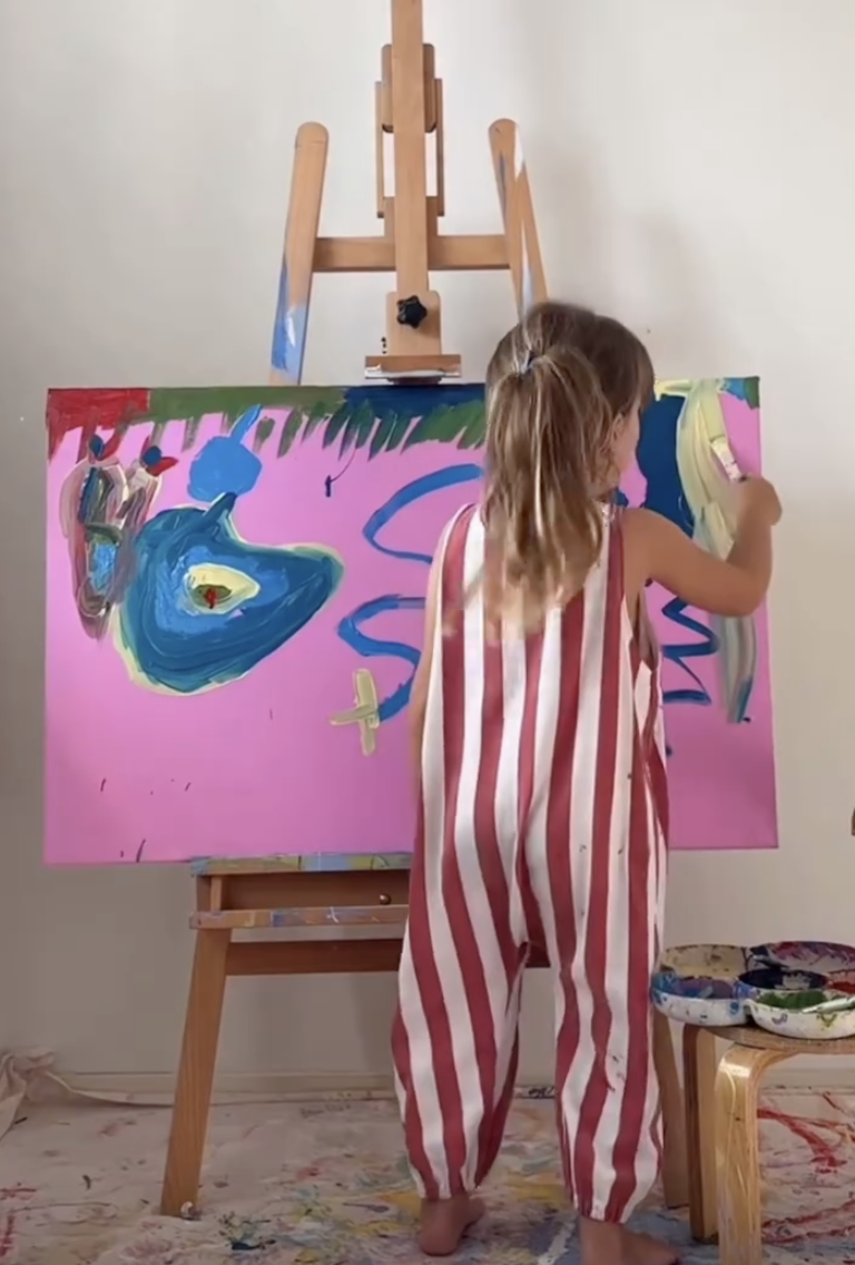 Child in red and white striped pajamas painting on a large pink canvas set on an easel.